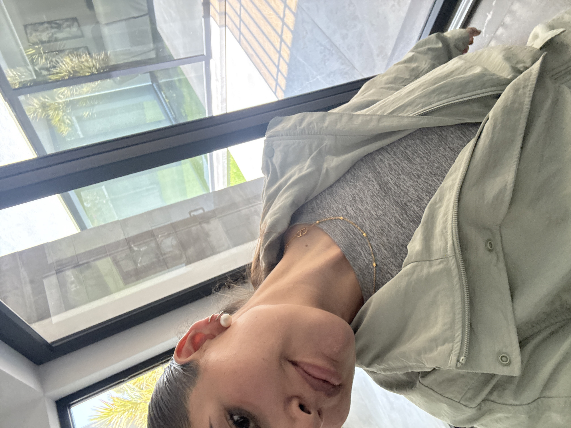 A partial view of a person, likely female, is captured in a close-up, angled portrait. The person's head and upper torso are visible, with their dark hair appearing pulled back. They are wearing a light grey, possibly ribbed, top, layered with an open, light green or khaki utility-style jacket. A delicate gold chain necklace with a small pendant is visible around their neck, and a pearl stud earring is prominent in their left ear, accompanied by a smaller hoop earring higher on the lobe. The person looks towards the camera with a neutral to slightly pensive expression.
The background is dominated by a large glass ceiling or roof structure with dark frames, suggesting a modern architectural design. Through and reflected in the glass, bright outdoor elements are visible. These include lush green foliage, resembling palm fronds or similar plants, a section of a green lawn, and reflections of surrounding architectural details, possibly from an adjacent building or patio. The bright, clear natural light indicates it is daytime, likely sunny. The setting appears to be a well-lit indoor or semi-outdoor space, such as a sunroom, atrium, or a room with extensive skylights, located in San Bernardino Tlaxcalancingo, Mexico. There is no visible text in the image.