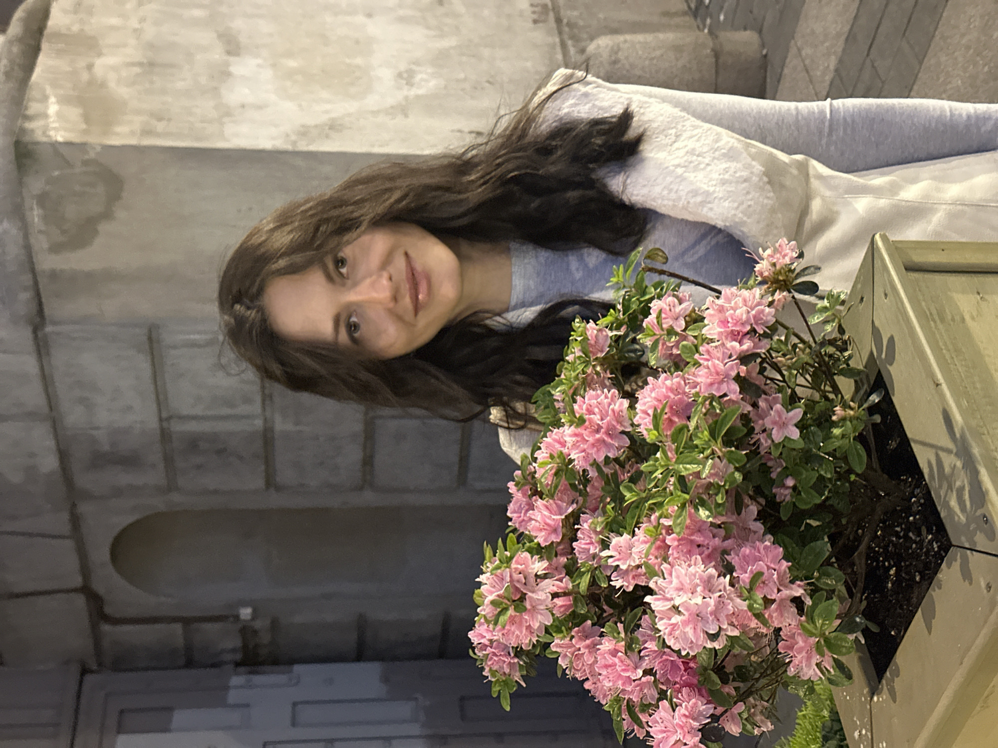 An evening or night-time portrait-style photograph shows a young woman next to a blooming plant in an urban setting, likely in Saint Petersburg, Russia. The scene is illuminated by artificial light, casting soft shadows.
The main subject, positioned on the left, is a woman with long, dark, wavy hair. She is looking directly at the viewer with a gentle, pleasant smile, her head slightly tilted. She wears a light-colored, possibly off-white or very light grey, textured jacket or cardigan over a light blue t-shirt. Her features are soft, and her eyes are dark.
To the right of the woman, and slightly in the foreground, is a wooden planter box filled with a lush plant bearing numerous small, light pink flowers, characteristic of an azalea bush. The flowers are delicate and densely packed, surrounded by green foliage. The planter box appears to be painted a muted, light green or greyish-brown color.
The background consists of large, rectangular blocks of what appears to be stone or concrete, forming a wall or building facade. The grey tones of the architecture suggest an urban environment. Some areas of the background show subtle variations in texture or possible water stains, indicating weathering. There is no visible text in the image. The overall mood is serene, with a focus on the woman and the vibrant flowers against the muted urban backdrop.