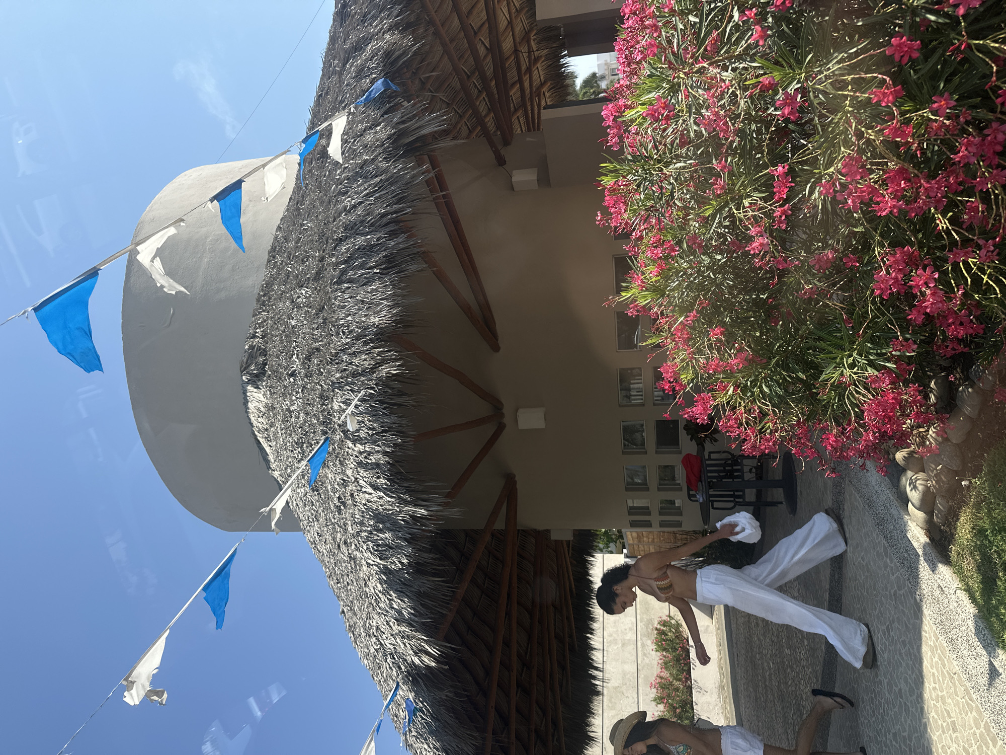 The image shows a brightly lit outdoor scene in Acapulco de Juárez, Mexico. A circular structure with a thatched roof dominates the left side of the frame. White and blue triangular pennants are strung along the perimeter of the roof. Adjacent to the structure, on the right, is a building with a tan exterior and multiple small, square windows. A large flowering bush with magenta blossoms is beside the building.
Two women are visible on a paved area in the foreground. One woman wears a bikini top and white pants, holding a white garment. Another woman is near the bottom of the frame, lying on the ground and wearing a sun hat. Outdoor furniture is visible in the background, including a table and chairs.