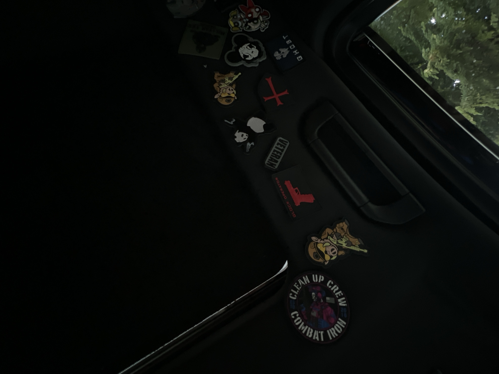 The image captures a dark interior of a vehicle, focusing on the headliner and a collection of morale patches affixed to it. The scene is illuminated dimly, suggesting nighttime.
On the dark gray headliner, a variety of colorful and monochrome patches are displayed. Visible patches include:
- A black patch with white text "GHOST" and a skull graphic.
- A red inverted cross on a black patch.
- A black rectangular patch with white text "VETERAN".
- A black patch with a red silhouette of a handgun and smaller white text that appears to read "BLOCK OPERATION" or similar.
- A circular patch with "CLEAN UP CREW" inscribed along the top and "COMBAT IRON" along the bottom, surrounding a stylized, colorful character that resembles a cyborg or armored figure.
- Several cartoon-style patches, including one featuring a character resembling Mickey Mouse with a distressed, skull-like face, another resembling Minnie Mouse with a skeletal or robotic design, and a character resembling a pig or boar in tactical gear holding a rifle.
- A white panda-like character patch.
- A partially visible green patch in the upper left corner.
To the upper right, a sunroof is open, revealing a view of dark green tree foliage outside, subtly illuminated by an unseen light source. A dark handle or grab bar is also visible on the headliner. The overall lighting implies a dark environment, likely inside a car at night. No people are visible, and no specific activity is taking place.
