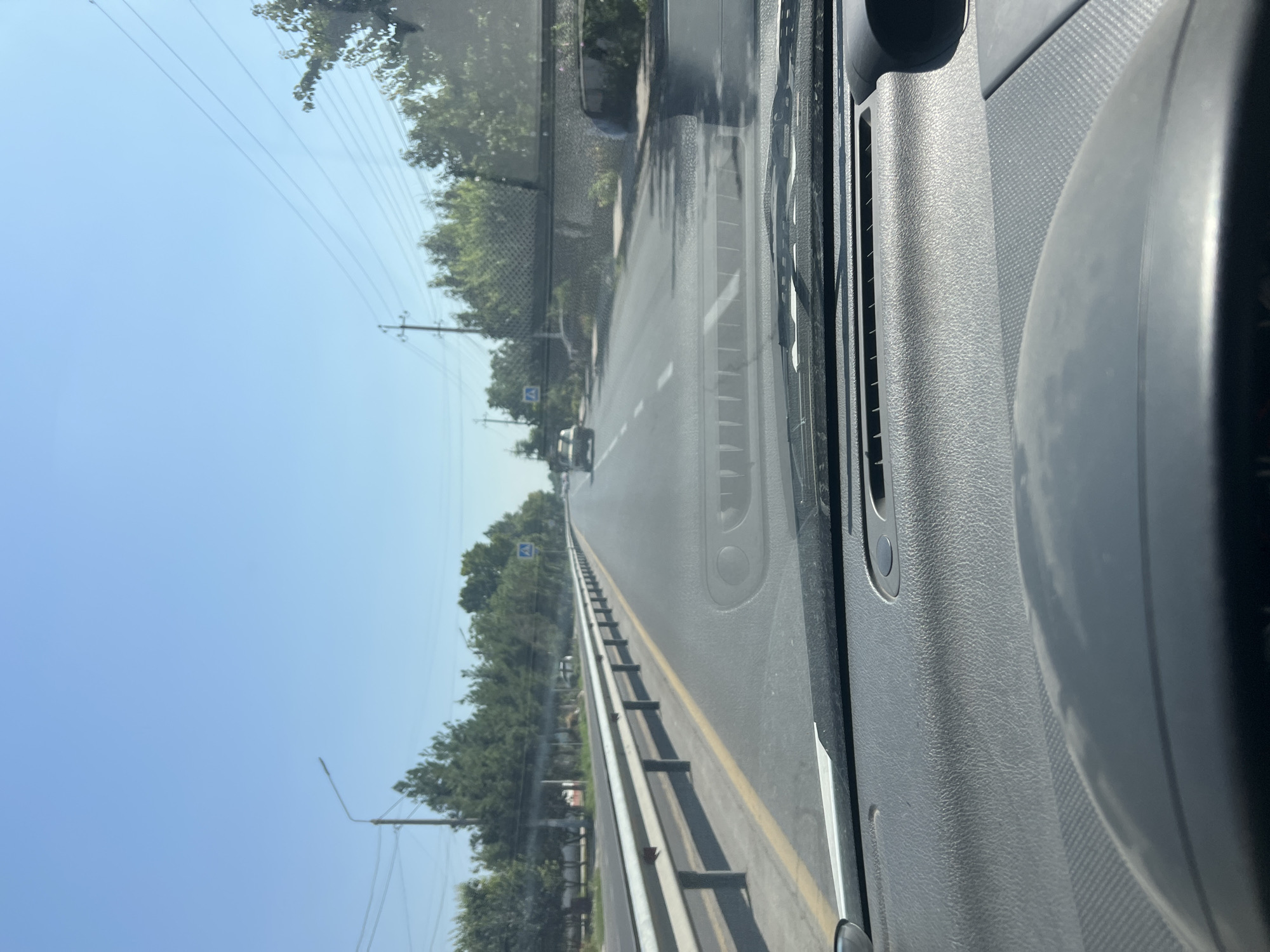 A first-person perspective shot from inside a vehicle captures a straight, paved road with a clear blue sky overhead. Trees line the right side of the road, and a guardrail is visible on the left. The road is marked with a yellow line on the left and white dashed lines in the center. A pedestrian crossing sign is posted on the right side of the road. Two vehicles are visible on the road ahead, one in the distance and another closer. The dashboard of the vehicle in the foreground is visible, with a textured gray surface and a speaker grille. The image was taken in Uymavut, Uzbekistan.