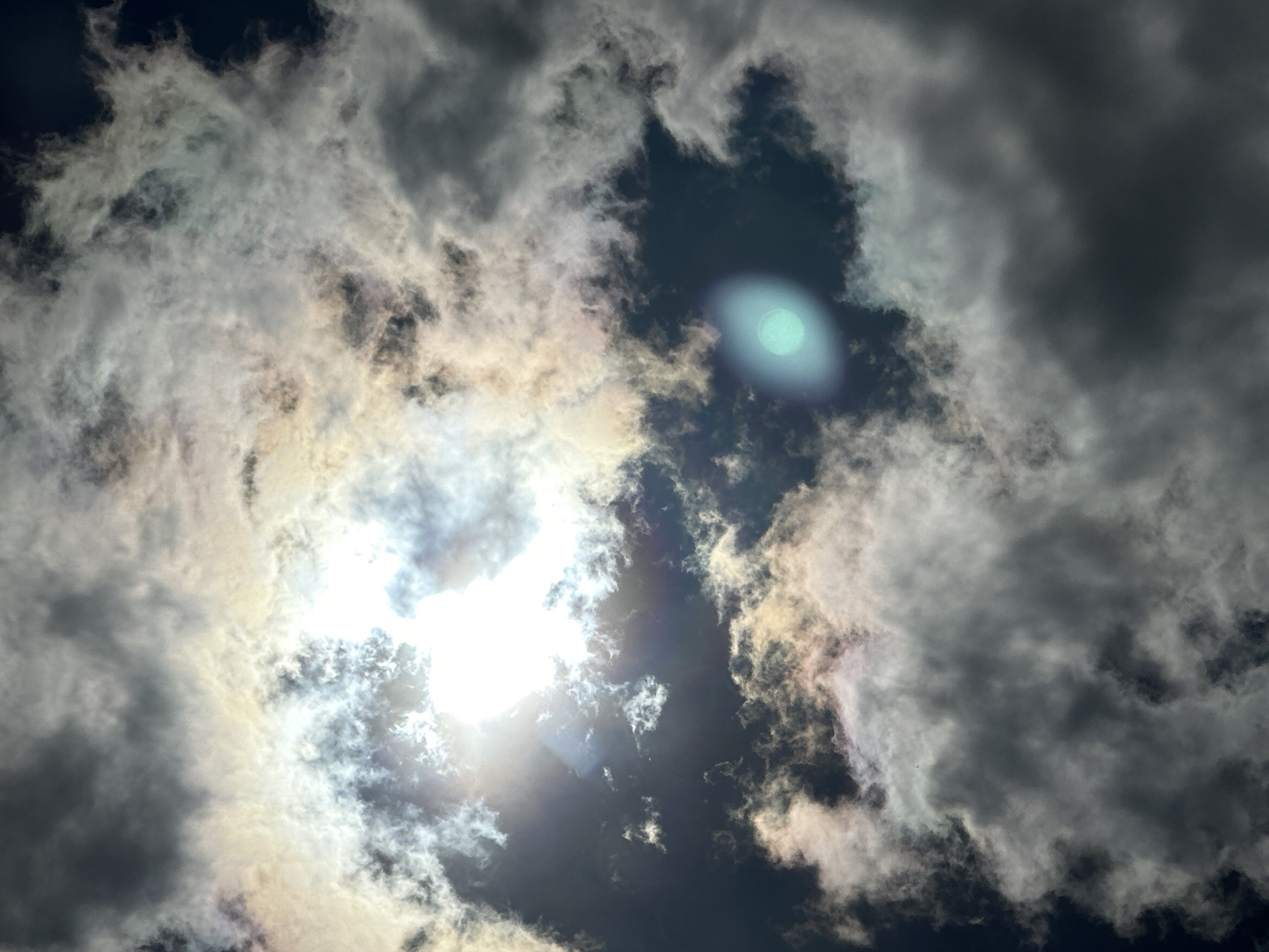 The image is an extreme close-up of a sky filled with dramatic clouds. The sun is partially obscured by these clouds, creating a bright, blown-out area in the upper center of the frame. Around the sun, there are iridescent or rainbow-like effects within the clouds, suggesting the presence of light refraction. The clouds themselves vary in density and color, ranging from dark, ominous grays to bright whites and yellowish-whites where they are illuminated by the sun. In the lower right quadrant of the image, a distinct, soft, circular glow of greenish-blue light is visible, possibly an artifact or a lens flare. The background sky, where not obscured by clouds, is a deep, dark blue. There are no discernible objects, people, or any context to suggest a specific location within a city or any environmental details beyond the sky and clouds.