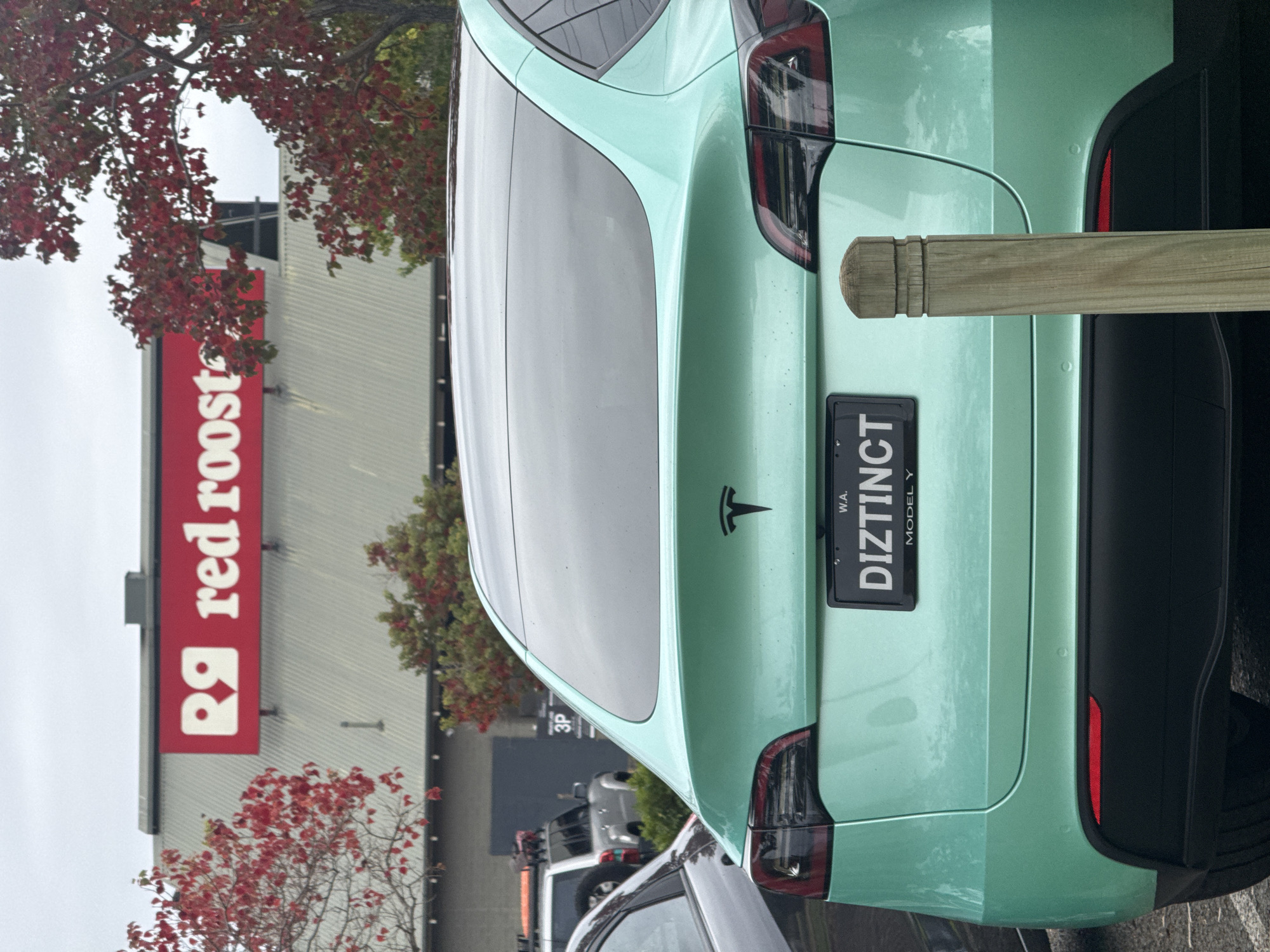 A light green Tesla Model Y, with the license plate "DIZTINCT," is parked in front of a Red Rooster restaurant in Bassendean, Western Australia. The car is positioned near a wooden post or barrier. Red-leaved trees are visible in the foreground and surrounding the building. Other vehicles are partially visible in the background, further down the street. The sky is overcast.