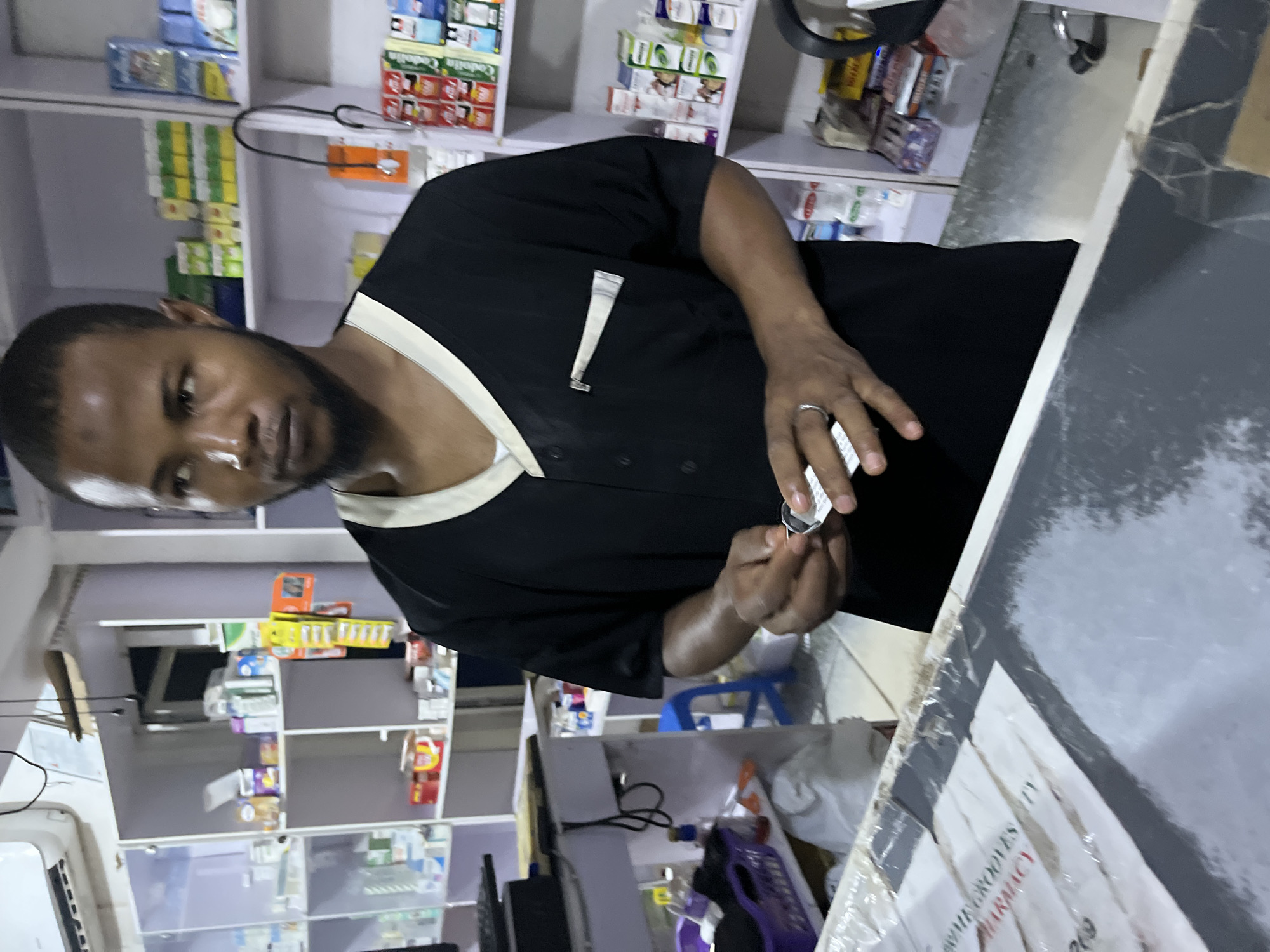 An individual is in a pharmacy. The individual is a dark-skinned male wearing a black shirt with white trim around the collar and a metal pen clip on the chest. The individual is holding a small white object. Shelves stocked with pharmaceutical products and medication are visible in the background. The location is Abuja, Nigeria based on the text "OME GROOVES PHARMACY" visible on the counter.