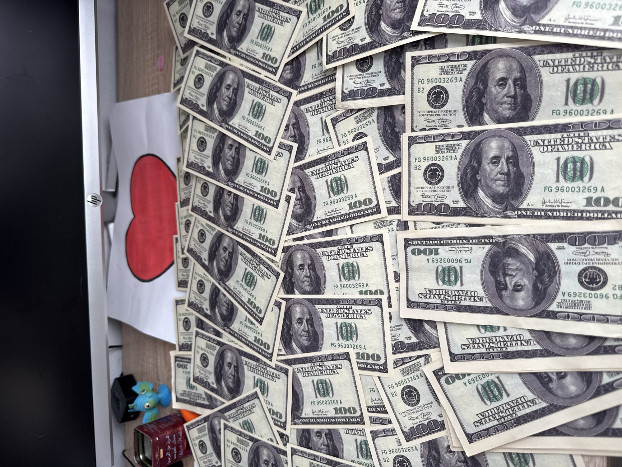 The image presents a desk or countertop cluttered with multiple 100 US dollar bills. The bills are primarily arranged in a haphazard fashion, covering a large portion of the surface. In the background, partially obscured by the money, there's a white piece of paper with a crudely drawn red heart on it. To the left, there's a visible part of a computer monitor. On the surface near the monitor are some small figurines and a box with Arabic writing. The location is not specified, but it is cluttered.