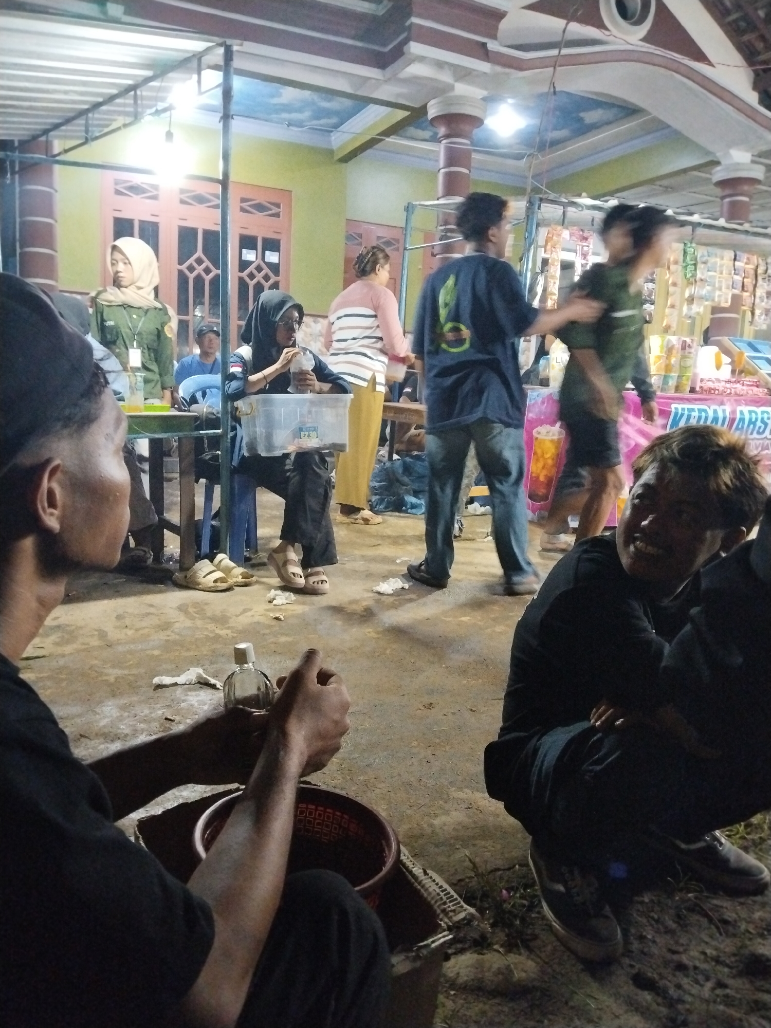This media file captures a lively evening or nighttime gathering in Jenggrik, Indonesia, likely at a communal area or food stall. Artificial lighting illuminates the scene under a covered structure with decorative pillars and windows in the background. The ground appears to be a mix of dirt and concrete.
Numerous individuals are present. In the immediate foreground, a man, seen from behind, is focused on a small clear bottle and a red plastic basket. To his right, another man squats, looking towards the viewer with a broad smile. In the mid-ground, a woman in a black hijab and glasses sits at a table, holding a clear plastic container with a straw. Next to her, a woman in a light-colored hijab and a green uniform stands, possibly bearing a badge with "UTM" visible. A man in a blue shirt and cap is partially visible seated nearby. Further back, two men appear in motion, one in a dark blue shirt and another in a green shirt, adding to the dynamic atmosphere. A woman in a striped top is seen from behind on the right.
The activity suggests a social event, with people eating, drinking, moving around, and interacting. The man in the foreground right displays a cheerful emotion. Notable objects include the small bottle, basket, clear drink container marked "FZ," a table, chairs, and shelves holding various merchandise in the background. A banner or poster in the background features an image of a drink and partial text "KEDAI ARSA," likely indicating "Arsa's Stall." The scene radiates a casual, friendly ambiance typical of an evening gathering in a community setting.