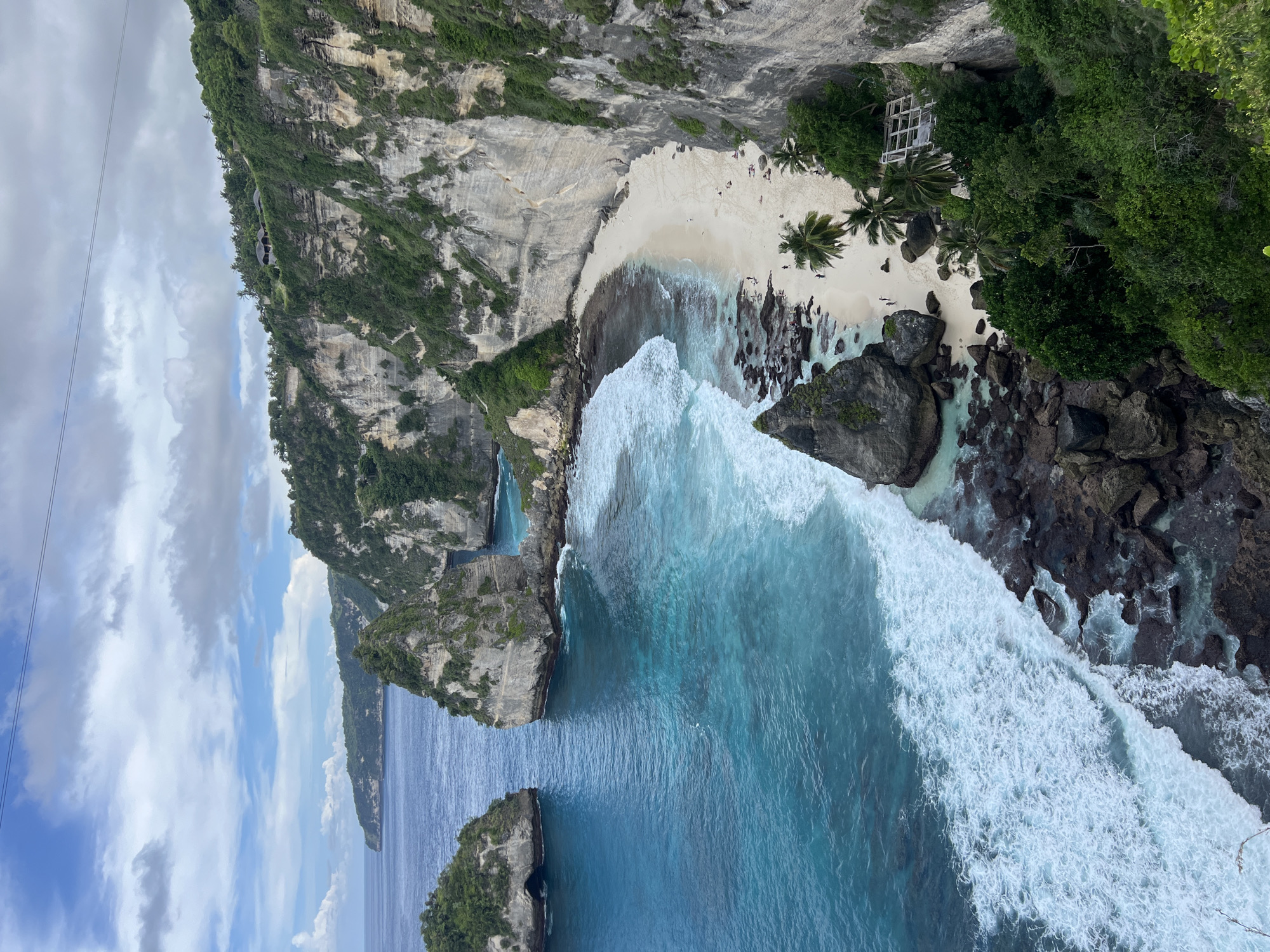 An aerial, panoramic view captures the stunning Diamond Beach in Nusa Penida, Indonesia, nestled between majestic, vegetation-covered cliffs and the vibrant turquoise ocean. The scene features a crescent-shaped stretch of white sandy beach at the base of towering, grey and beige limestone cliffs.
The crystal-clear water transitions from a light turquoise near the shore to a deep blue further out, with powerful white-capped waves continuously breaking onto the rocky shoreline. Numerous dark, jagged rock formations are scattered along the water's edge and partially submerged, adding texture to the coast.
On the white sand, several small figures of people are visible, some walking near the water and others relaxing on the beach. Palm trees grow on the beach, providing shade, and dense green foliage blankets the steep cliffs. A distinctive white, open-sided structure with a gabled roof is nestled among the trees on the beach, while smaller, dark-roofed structures are faintly visible on the cliff tops in the distance.
The sky above is bright with scattered white clouds, suggesting a clear day, likely morning or early afternoon, given the strong natural light. Two thin parallel wires, possibly from a zipline or cable car, cut across the upper left portion of the sky. The overall impression is one of breathtaking natural beauty and a serene, secluded paradise. No visible text is present in the image.