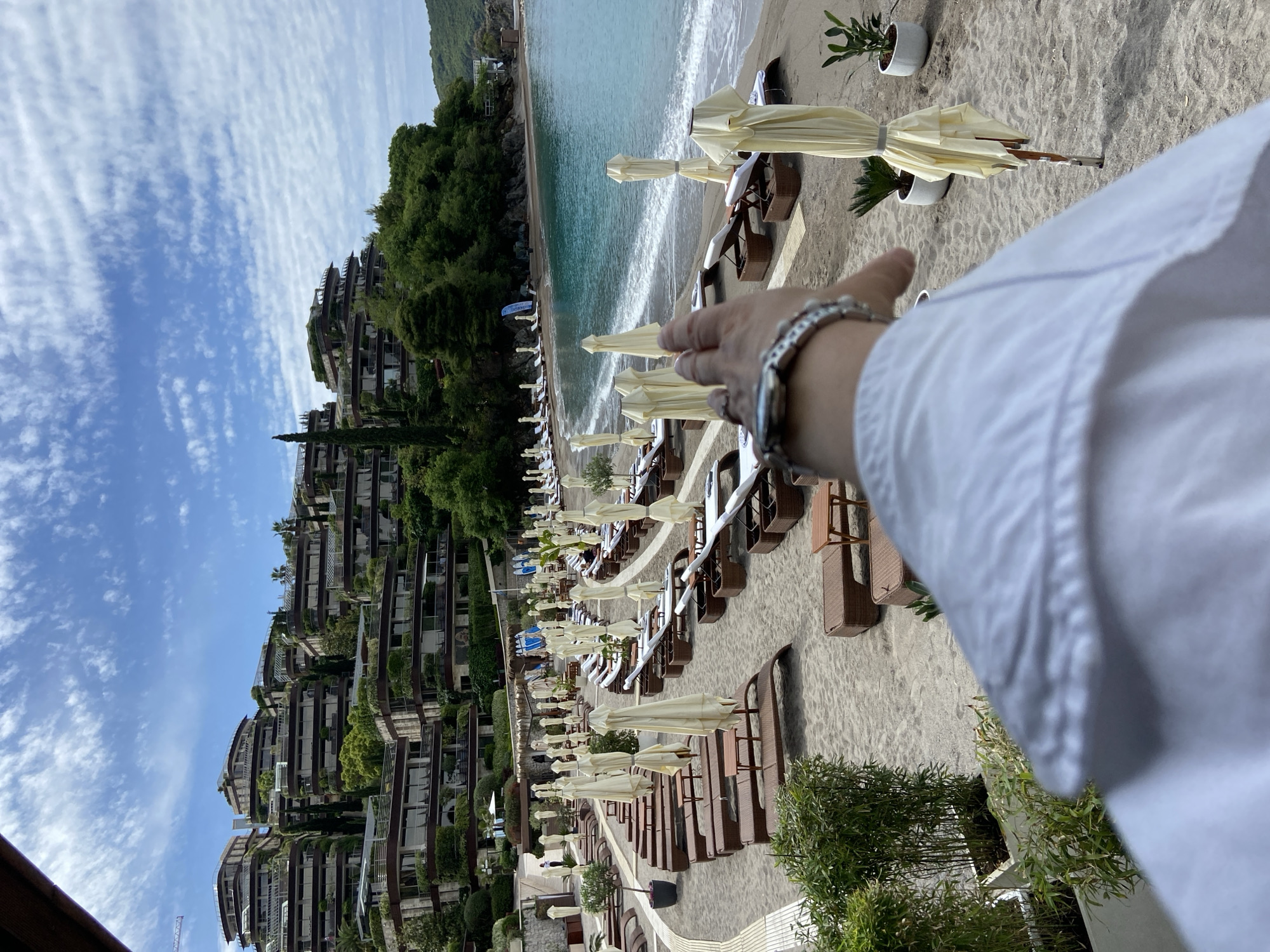 The image depicts a beach scene in Budva, Montenegro. The focus is on rows of beach loungers with beige umbrellas arranged along a sandy beach. In the background, there is a multi-tiered building complex covered in greenery, partially obscured by the blue sky with scattered clouds. A clear, turquoise sea meets the shore. In the foreground, a person's hand and part of a white shirt sleeve are visible. The bracelet on the wrist is a silver linked bracelet. The beach scene suggests a resort or private beach area.