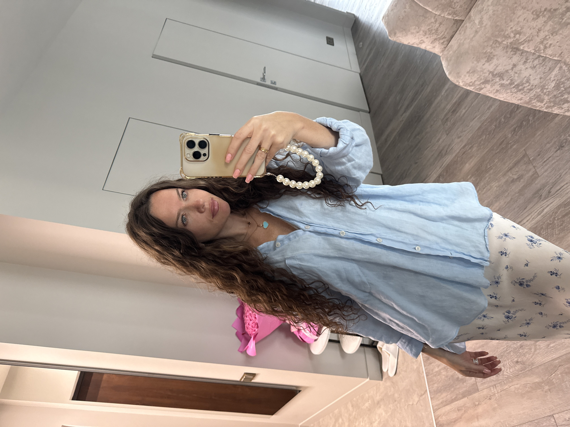 A woman with long, curly brown hair is taking a mirror selfie in what appears to be an apartment hallway in Khimki, Russia. She is wearing a light blue linen button-down shirt and a light-colored skirt with a floral pattern. She has a turquoise pendant necklace and a ring on her right hand. Her phone, with a gold case and a pearl wrist strap, is held up to take the picture. In the background, there is a large mirror, a doorway, and a beige upholstered couch. To her left, a bouquet of pink roses is visible on a small table. The floor is a light wood laminate.