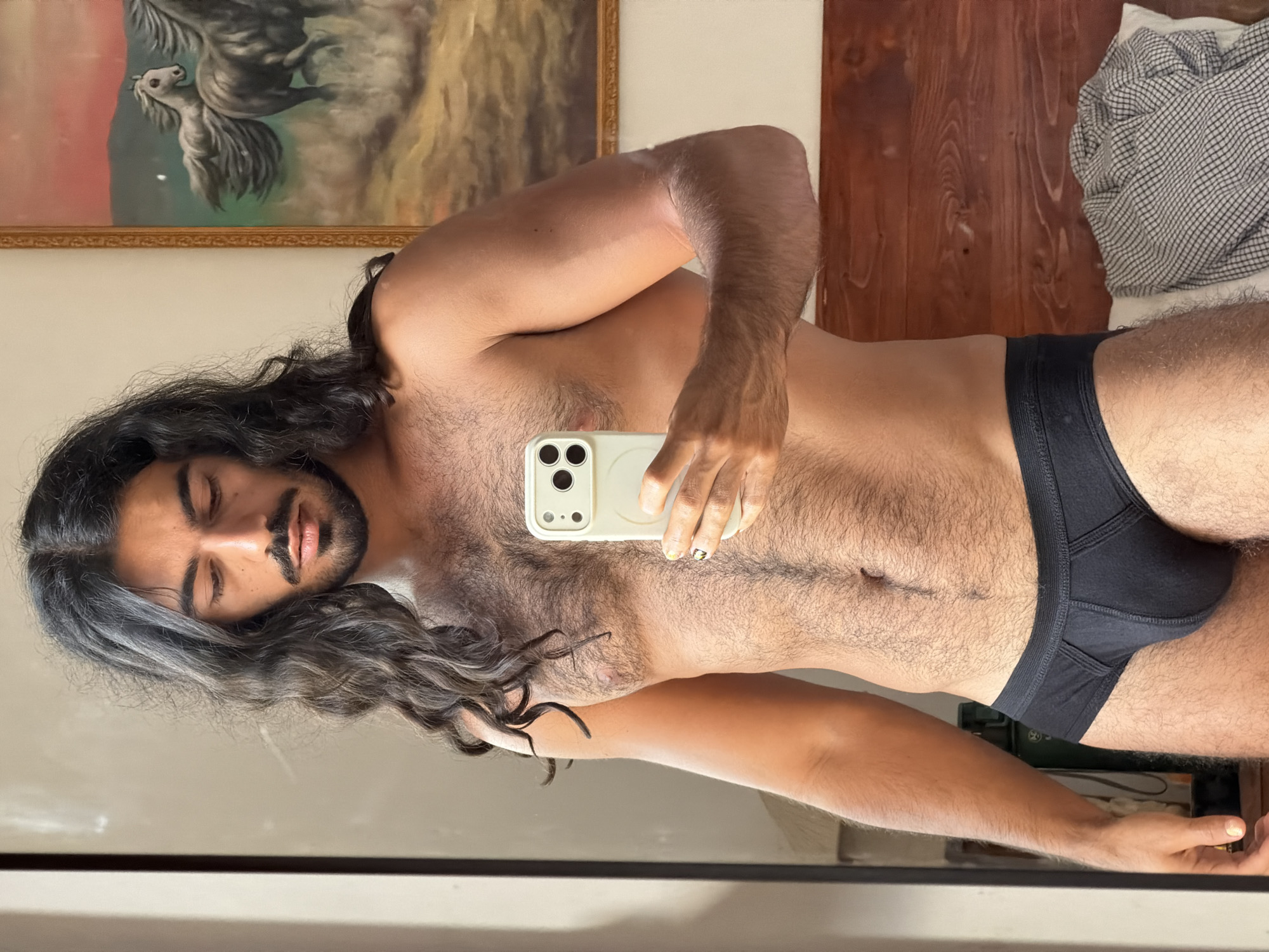 A shirtless man with long, dark, wavy hair is taking a selfie in a mirror. He is wearing black briefs. In the background, a painting of horses and a wooden headboard are visible. The lighting suggests it is daytime. The location is North Kuta, Indonesia.