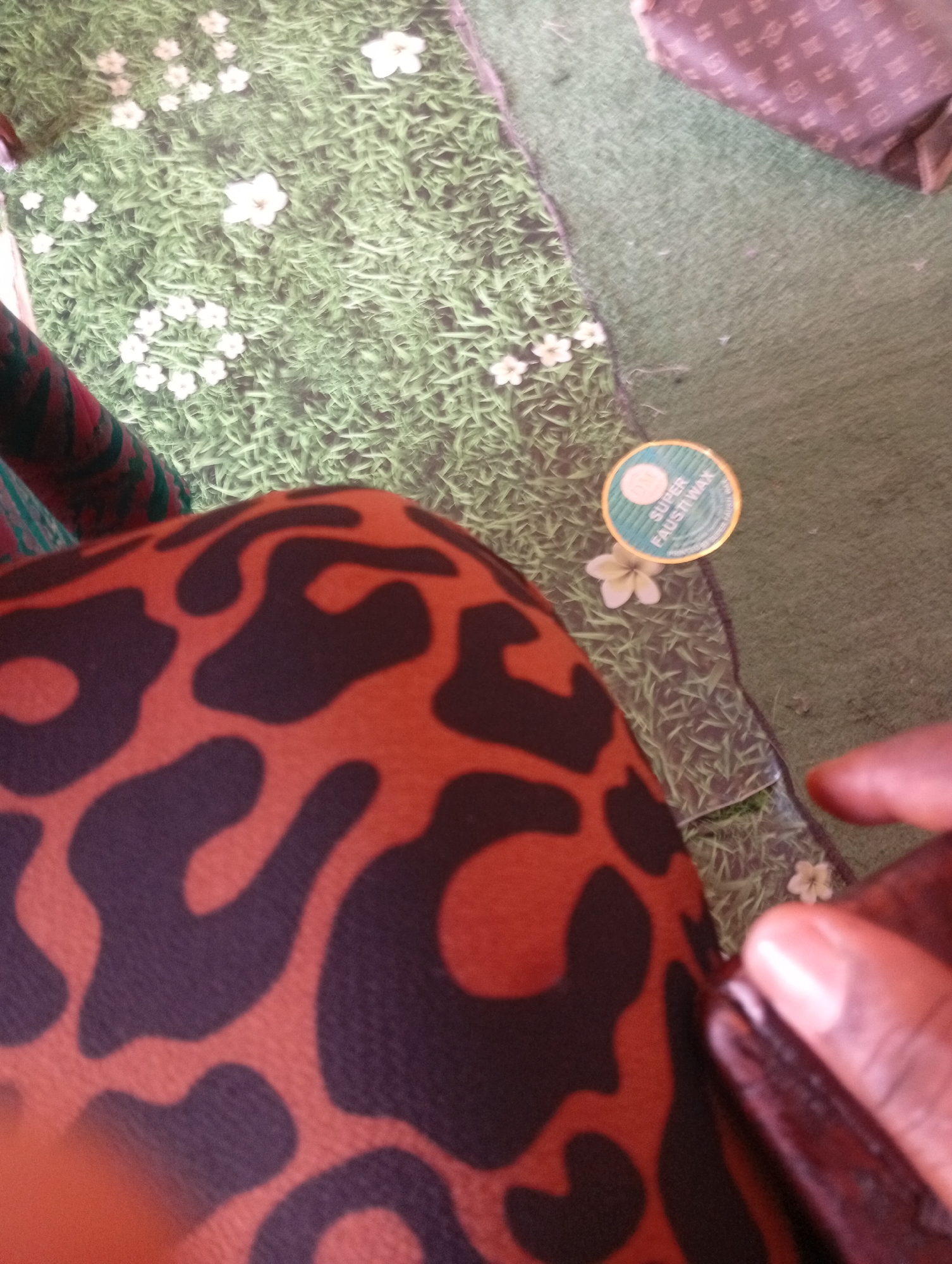 A close-up shot shows a person's lap covered in a reddish-brown fabric with a black leopard-print pattern. The fabric appears to be part of clothing or upholstery. To the right, a blurred hand with dark skin is visible, with fingers extended towards the lower right corner.
The background features a surface that resembles artificial grass with white flowers scattered across it, creating a somewhat whimsical or decorative scene. Partially visible in the upper right corner is a bag or piece of luggage with a repeating pattern of brown squares and diamond shapes.
On the artificial grass, a small, circular, gold-rimmed sticker with a light blue center is prominently displayed. Text is visible on the sticker, appearing to read "SUPER FAUSTI WAX". A single white flower is positioned just below the sticker. The lighting suggests it is daytime. The overall impression is a casual, indoor setting with decorative elements.