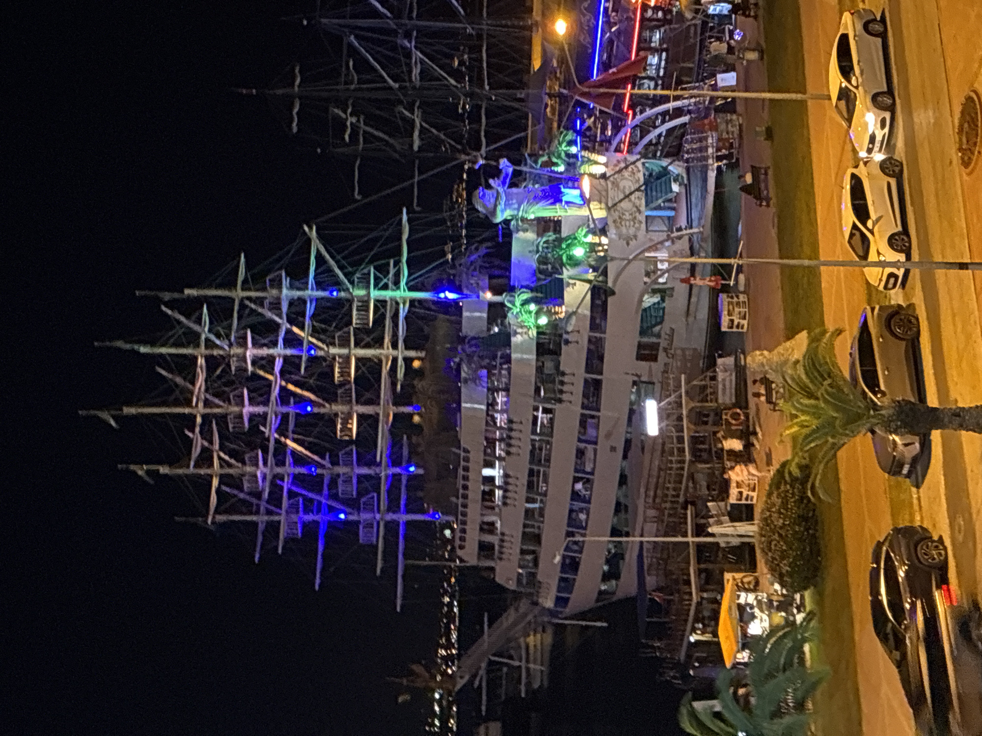 The image depicts a nighttime scene in Alanya, Turkey, featuring a large, illuminated pirate ship docked at a harbor. The ship is adorned with colorful lights, primarily blue and green, highlighting its multiple decks and masts. On the deck, a large statue of a muscular, bare-chested figure, possibly a mythological character or a pirate, is visible. Several people are seen on the ship's decks and in the surrounding area.
In the foreground, a street is visible with several cars parked. A dark-colored SUV is on the left, and two white cars, a hatchback and a sedan, are parked next to it. The street is illuminated by streetlights, casting a warm glow. Palm trees are present, with one on the right side of the image, its fronds partially obscuring the view.
Behind the pirate ship, another similar ship is partially visible, also with masts and rigging. In the distance, the cityscape of Alanya is visible, with lights dotting the landscape and extending towards the hills. The overall atmosphere suggests a lively tourist area, possibly a promenade or harborfront, at night.