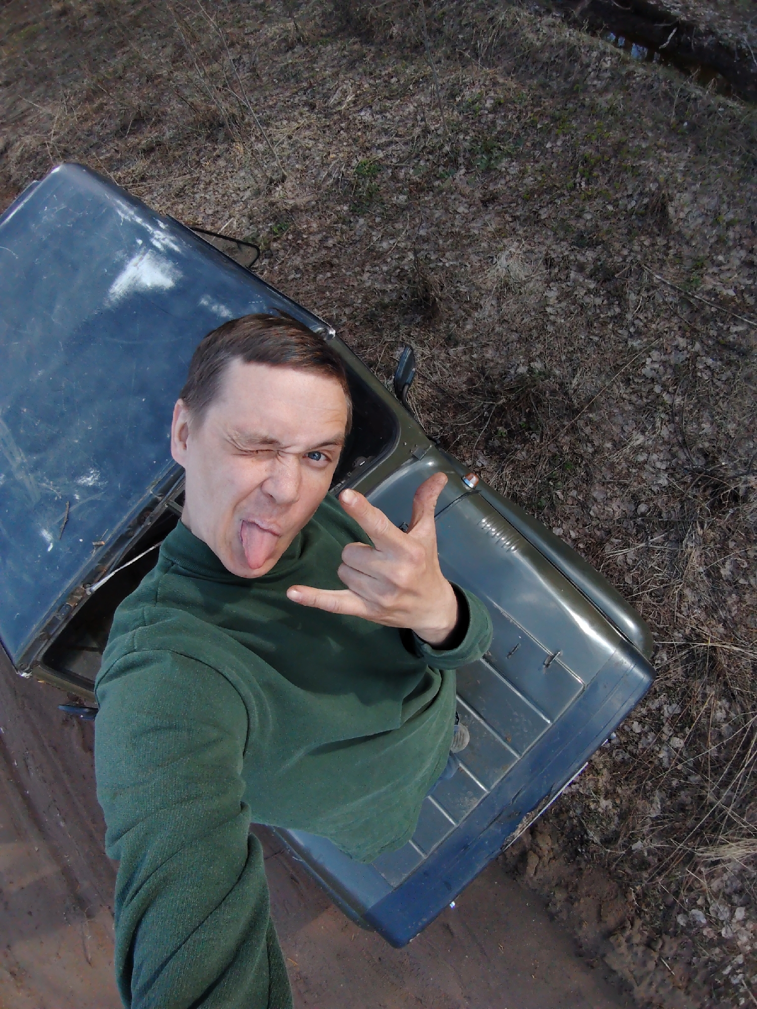 An overhead, high-angle selfie captures a man in Markovskii, Russia, playfully posing with his tongue extended and winking, while simultaneously making a "rock and roll" hand gesture with his left hand. He is wearing a dark green long-sleeved shirt, possibly a turtleneck. The man appears to be standing or leaning out of a dark-colored, rugged off-road vehicle, likely a jeep or similar utility vehicle, which shows signs of use and minor damage on its surface.
The photograph is taken looking down on the man and the vehicle, which is situated on a dirt track or muddy path. The surrounding environment consists of natural ground covered extensively with dry, brown leaves and sparse, bare branches, indicative of late autumn or early spring. The vegetation is largely dormant, suggesting a cool season. In the background, more of this dry, leafy terrain is visible, along with a hint of a small body of water or ditch to the far right.
The lighting is natural and appears to be daytime, possibly under an overcast sky or during early morning/late afternoon, as there are no harsh shadows. The man's expression conveys lightheartedness, mischief, and energy. There is no visible text in the image. The scene suggests an outdoor adventure or casual exploration in a rural or natural setting in Russia.