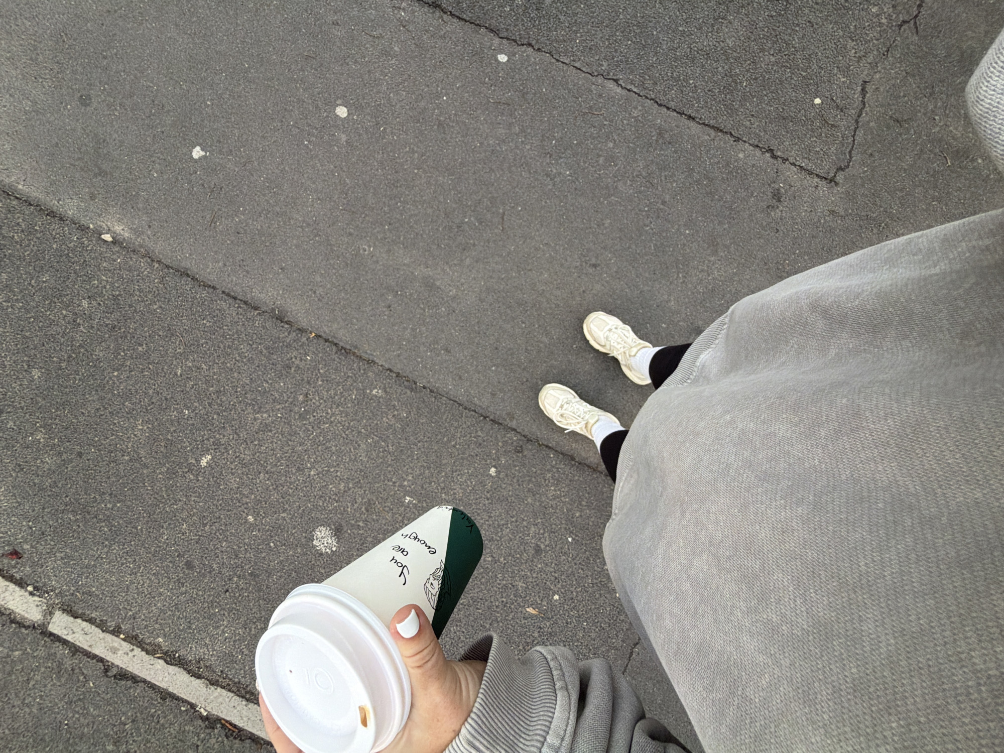 A high-angle, first-person perspective photograph captures a person's lower body and hand as they stand or walk on an asphalt surface in Cape Town, South Africa. The individual wears light grey, possibly denim or sweat-material pants, black socks, and pristine white athletic shoes. In their right hand, adorned with white nail polish, they firmly grasp a white disposable coffee cup with a white lid. The cup features a distinct dark green section around its top half and carries the uplifting message, "You are enough," written vertically, alongside an illustrated design that includes a stylized face or figure. The surrounding ground is dark grey, textured asphalt, showing minor cracks and scattered light-colored debris. The ambient lighting suggests it is daytime, likely under an overcast sky or in soft light, given the absence of harsh shadows.