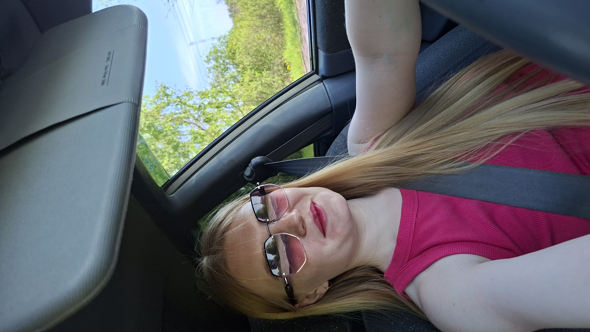 A young woman with long blonde hair and wearing sunglasses and a pink top is sitting in a car. She is wearing a seatbelt. The sun visor is down on the driver's side. Outside the window, there is greenery. The location is not discernible from the provided image.