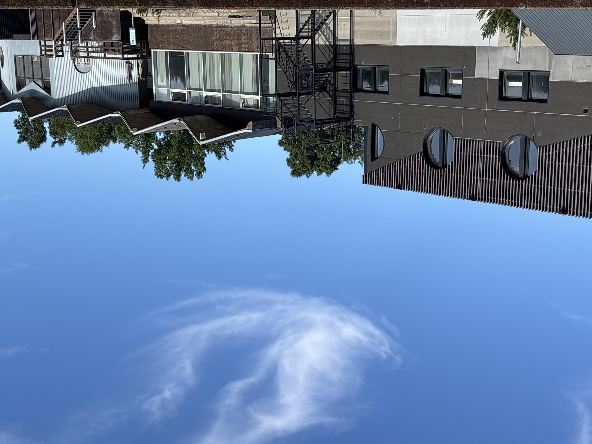 A modern building complex is depicted under a clear blue sky with a wispy cloud formation. The building on the left features a dark gray facade with vertical wooden slats at the top and three large circular windows. Below these, rectangular windows are visible. A dark gray metal staircase is attached to the side of this section. To the right, another part of the building has a wave-like roofline and large windows with white curtains. A wooden deck with railings and stairs is in front of this section. In the distance, a line of green trees stands against the sky. The scene appears to be in Haabneeme alevik, Estonia. There are no people visible in the image.