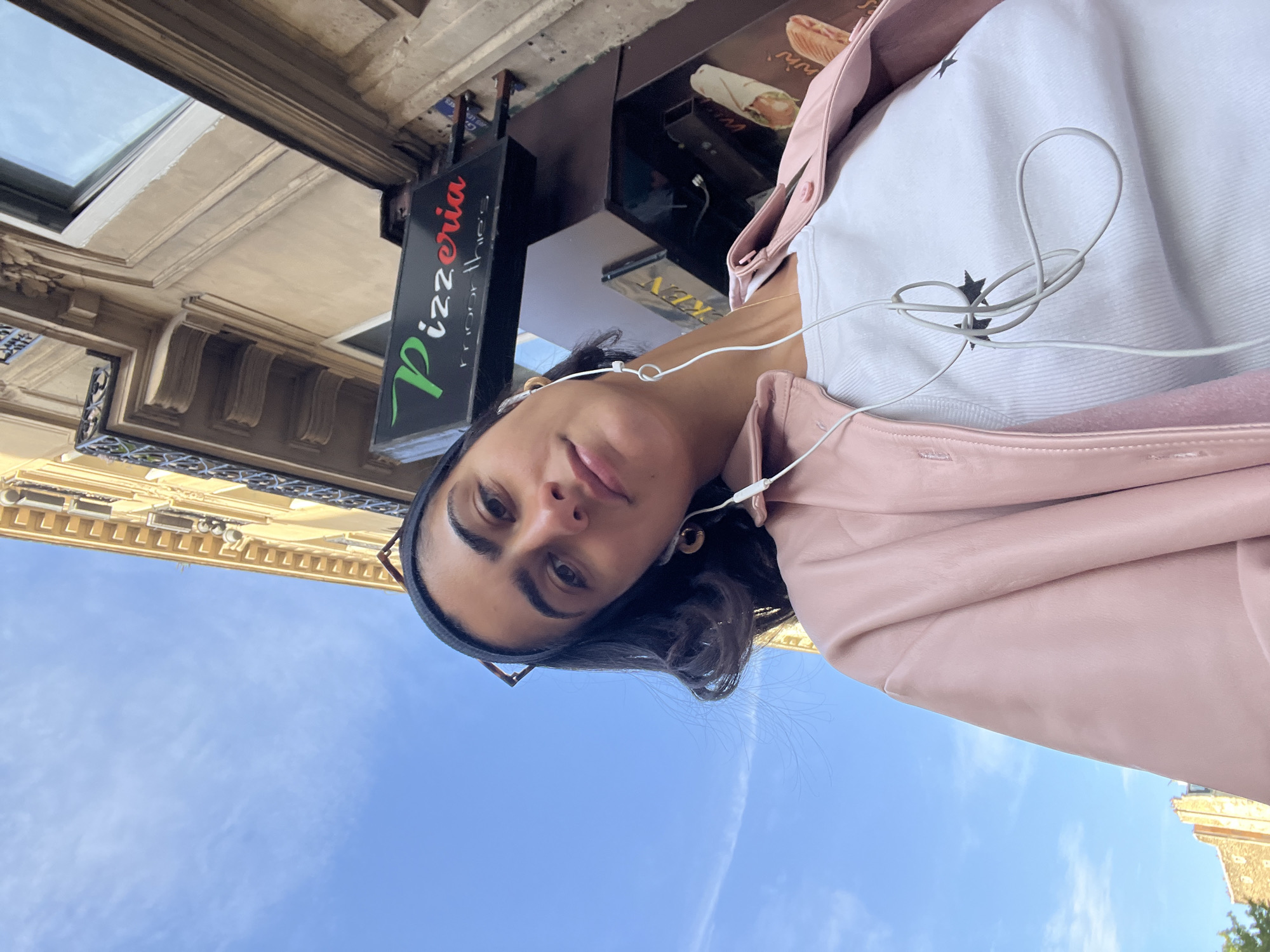 The image shows a woman with dark hair wearing a pink jacket and white top with headphones, standing in front of a building in Paris, France. A "Pizzeria" sign is visible in the background. The sky is blue with a contrail.