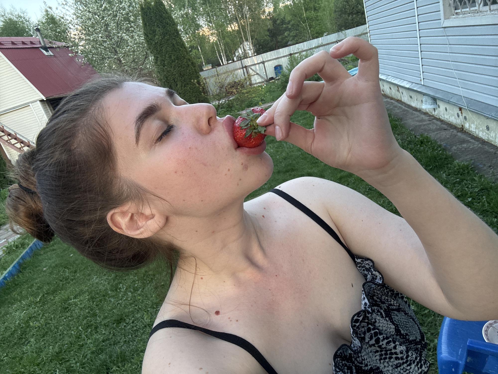 A woman is holding a strawberry to her lips in an outdoor setting, possibly a backyard in Мещёры, Russia. She has her eyes closed and appears to be about to take a bite. She is wearing a black and white print tank top. Her hair is pulled back in a bun. The background consists of a grassy lawn, a house with gray siding and a red roof, and trees. The image appears to have been taken at an angle, likely a selfie.