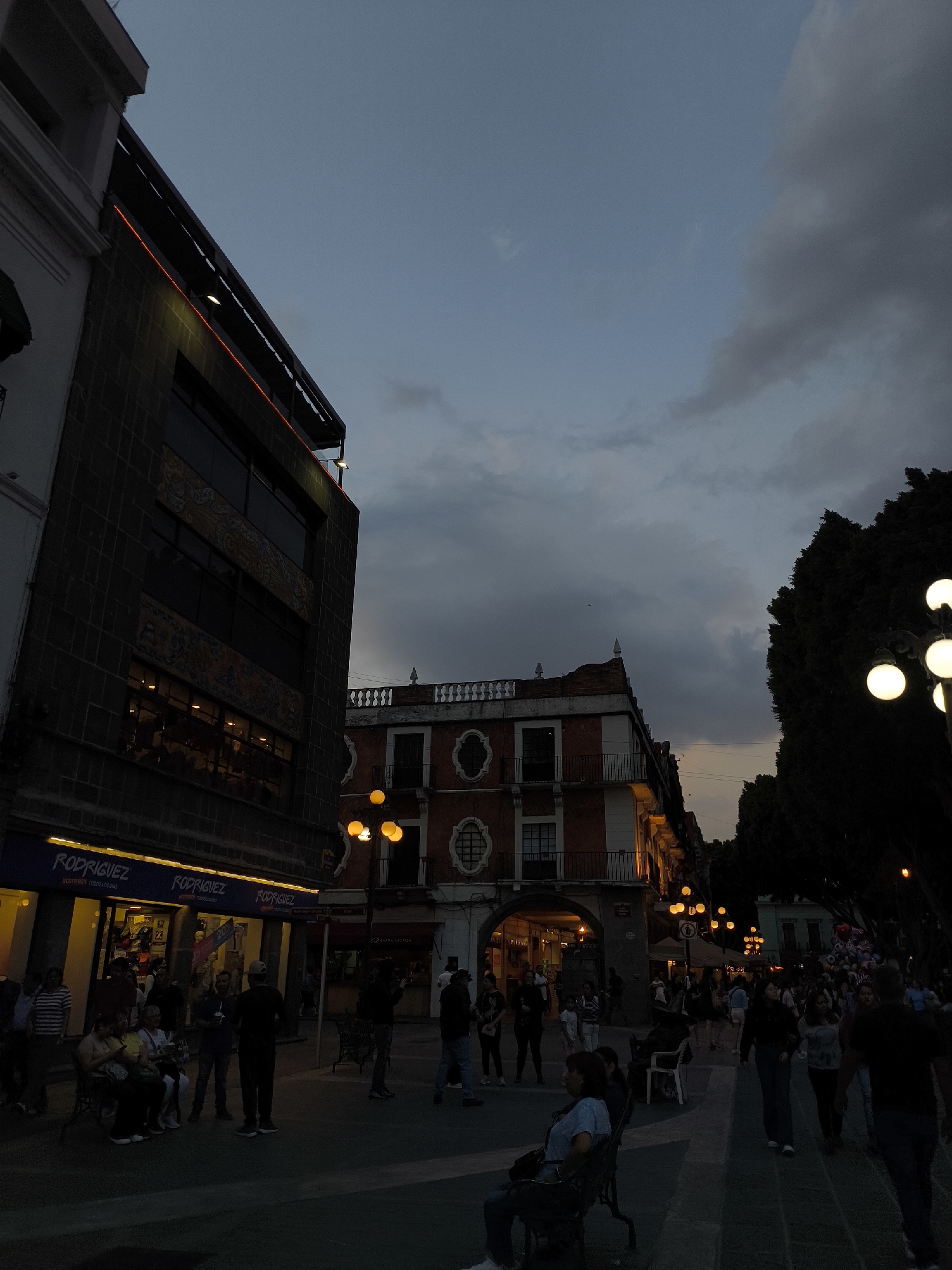 The image depicts a street scene in Puebla, Mexico, at dusk. Several people are present, some sitting on benches, while others walk along the pedestrianized area. Buildings line both sides of the street, with a "Rodriguez" store illuminated on the left. Streetlights are lit, providing additional illumination. The sky is cloudy.