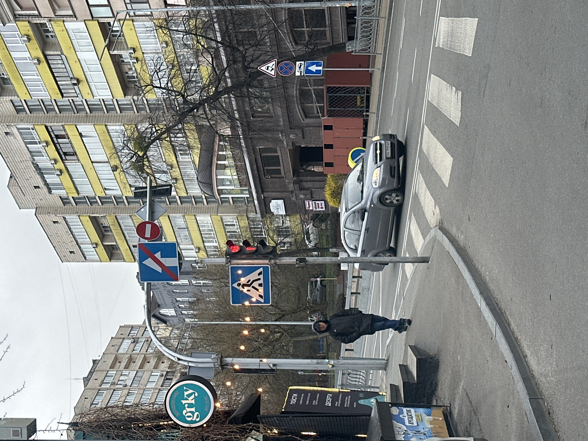 The image shows a street scene in Kyiv, Ukraine. A person is walking on the sidewalk near a crosswalk with a red traffic light illuminated. A gray car is stopped at the crosswalk. Street signs are visible, including a pedestrian crossing sign and a "no left turn" sign. Buildings with yellow and gray facades are in the background. There is a cafe with a sign "grky" visible.