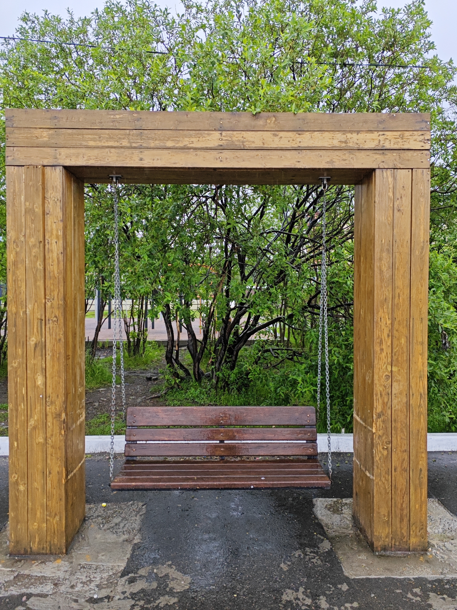 A wooden swing set with a brown wooden bench is set against a backdrop of lush green trees. The swing set is constructed from thick, polished wooden beams forming a rectangular frame. Two vertical posts support a horizontal beam from which the swing is suspended by silver chains. The swing bench itself is made of several horizontal wooden slats. The ground around the swing set is paved with a dark material that appears wet and reflective, suggesting recent rain. Some lighter-colored concrete or stone slabs are visible on the ground as well, particularly around the base of the wooden posts. The trees behind the swing are dense with vibrant green leaves, indicating a temperate season. The sky is overcast, contributing to the diffused lighting of the scene. The image is framed centrally on the swing set and bench, with the natural elements of trees and sky filling the upper portion. The location is identified as Polyarny, Russia. There are no people or animals visible in the image.