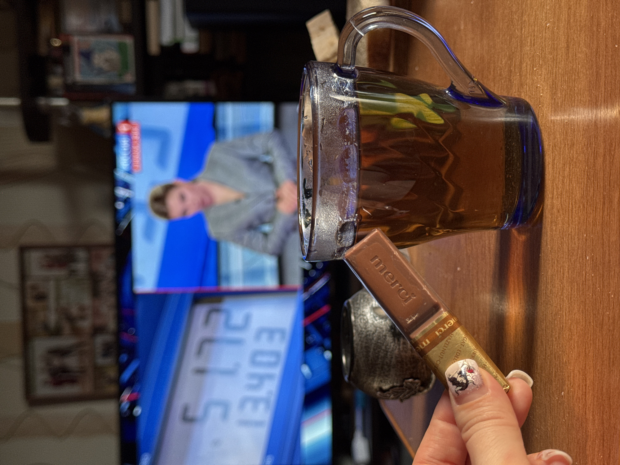 The scene depicts a partially filled blue glass mug of tea, with a chocolate bar labeled "merci" resting against it. A hand with decorated fingernails holds the chocolate. In the background, a television displays a news program featuring a woman in a gray outfit, with financial data visible below. The setting appears to be indoors, on a wooden surface. The location, Izhevsk, Russia, cannot be independently verified from the image.