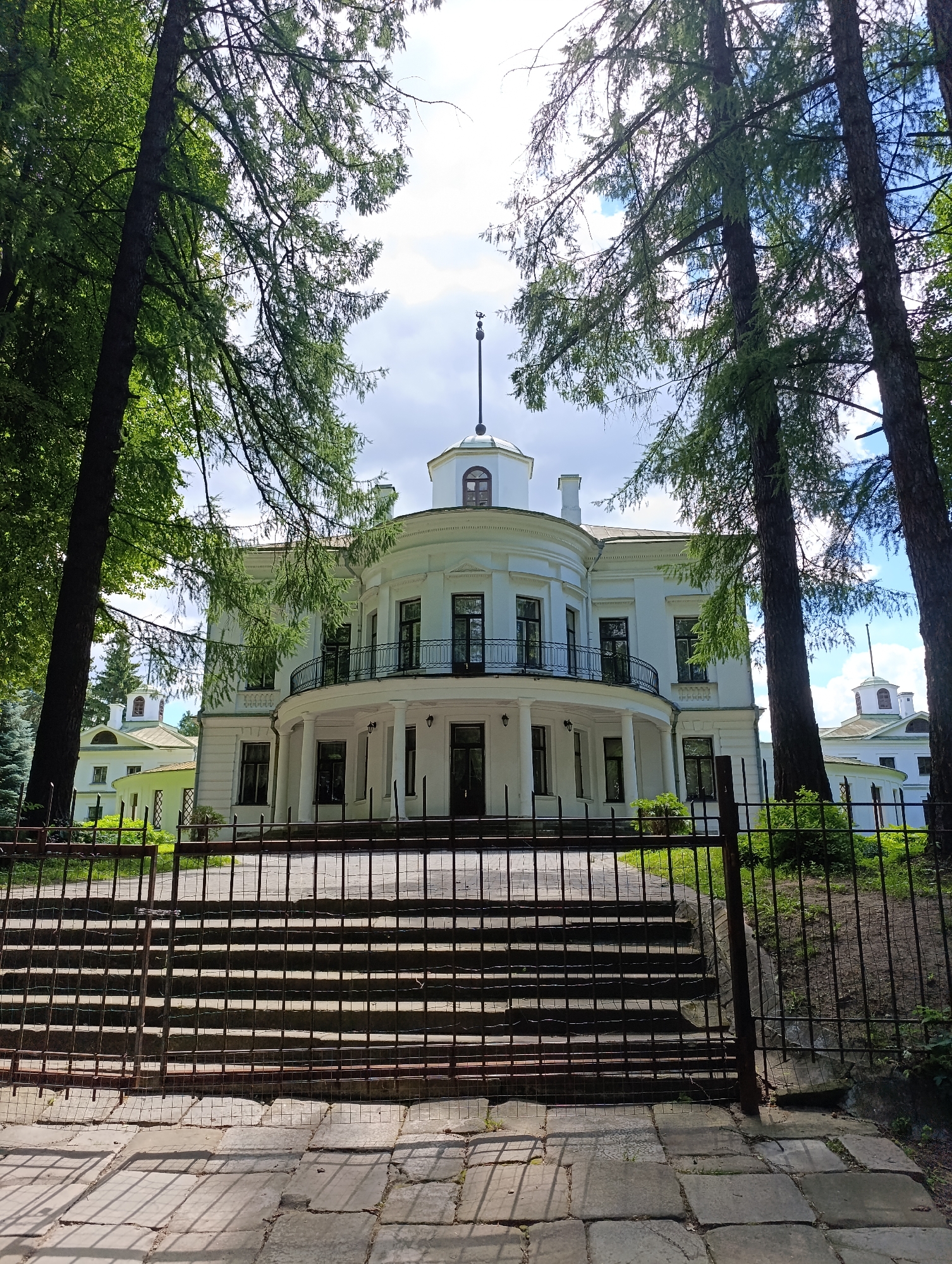 The image depicts a grand, white, neoclassical building with a rounded facade and a balcony supported by columns. Tall trees with lush green foliage flank the building, framing it within a natural, park-like setting. The sky is partly cloudy, suggesting it is daytime with fair weather. In the foreground, a dark metal fence with a gate, partially ajar, stands before a set of wide, stone steps leading up to the building. The steps are partially obscured by shadows cast by the fence and the surrounding trees, creating a dramatic play of light and shadow on the stone pavement. Behind the main building, a smaller, similarly styled structure with a domed roof is visible, hinting at a larger estate or complex. The overall impression is one of historical architecture immersed in a serene, verdant landscape, possibly a museum, a historical landmark, or a private estate. There is no visible text in the image. The scene evokes a sense of peace and grandeur, a moment captured on a bright, perhaps warm, day.
