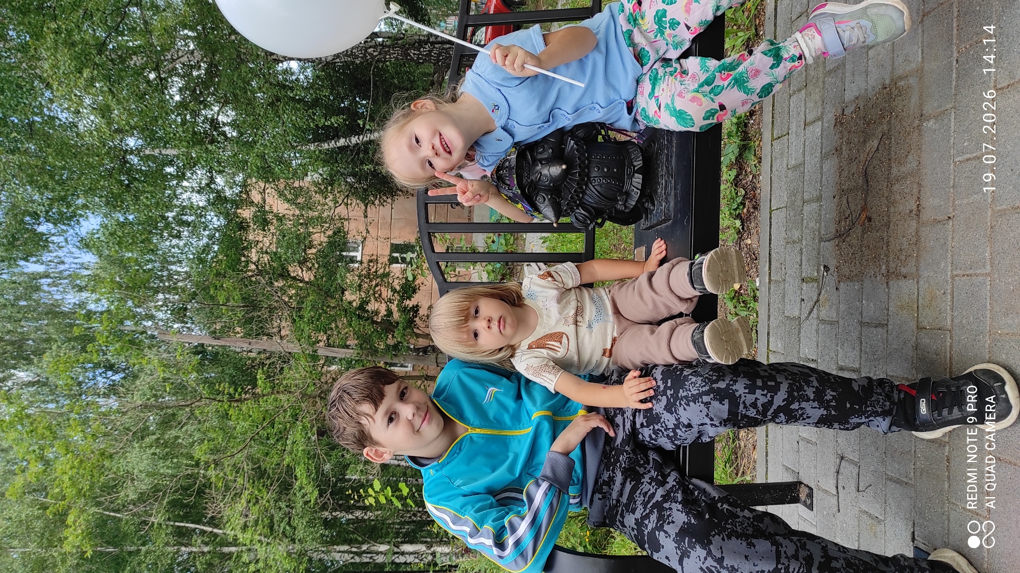 Three children are seated on a black metal bench in an outdoor park area. On the left is a boy with brown hair wearing a blue and yellow jacket and patterned pants. Next to him, in the center, is a younger child with blonde hair wearing a light-colored t-shirt with animal prints and beige pants. On the right is a girl with blonde hair, wearing a blue shirt and brightly patterned pants. She is holding a white balloon and making a peace sign with her fingers, looking at the camera. Between the two younger children is a dark-colored statue of a gnome-like figure. The background shows trees and a building. The ground in front of the bench is paved with grey tiles, with some dirt and debris visible. The timestamp "19.07.2026 14:14" and "REDMI NOTE 9 PRO AI QUAD CAMERA" are present in the lower corners of the image. This scene is located in the city of Chaikovsky, Russia.