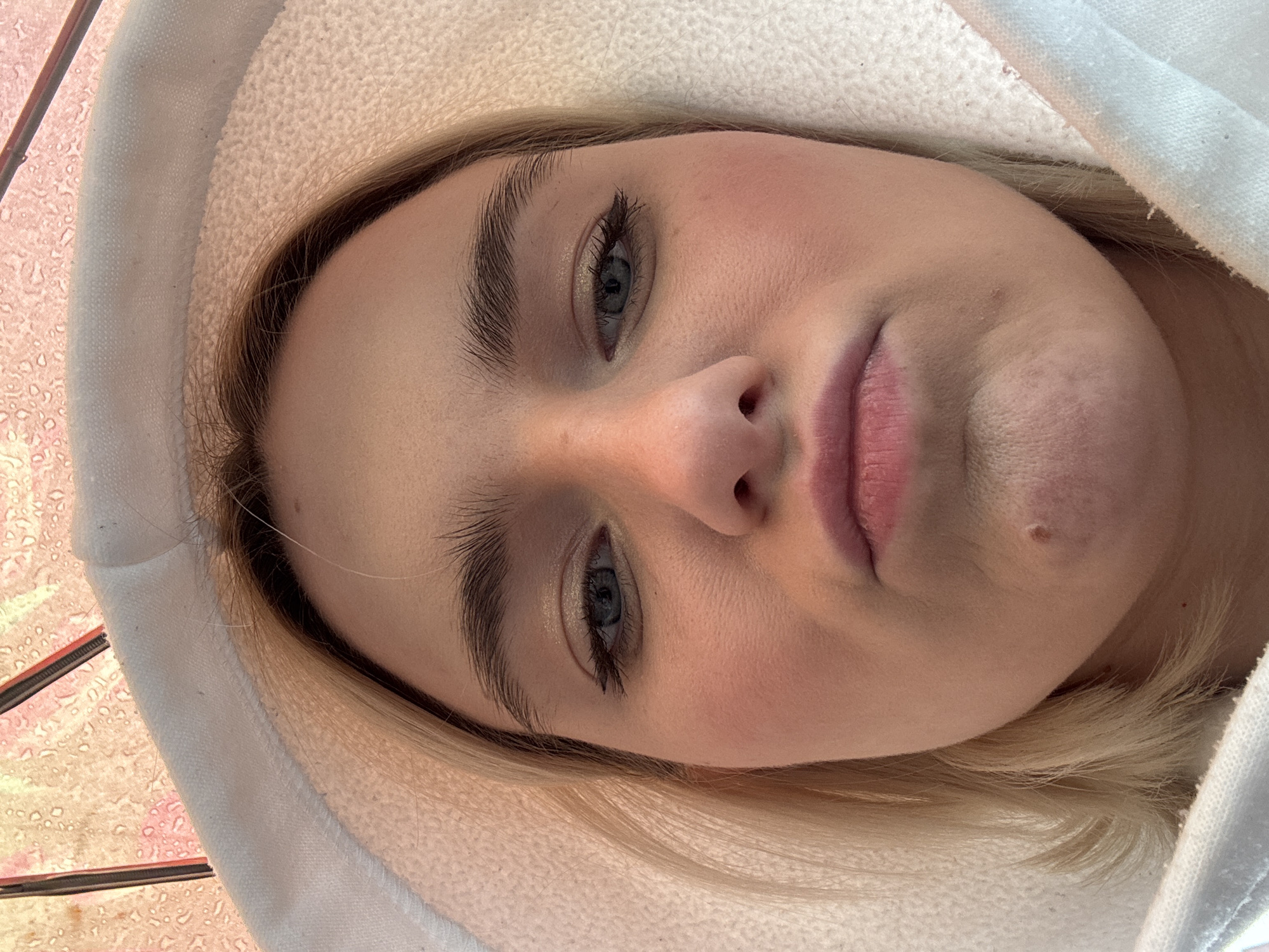 A close-up, upward-angled selfie of a fair-skinned person with light blonde hair, wearing a white hooded garment. The person has light blue eyes, prominent eyebrows, and pale pink lips. A small mole is visible on the right side of their chin. The background is indistinct but appears to include a blurry pattern suggesting light and color. The location in Saint Petersburg, Russia, cannot be determined from the image.