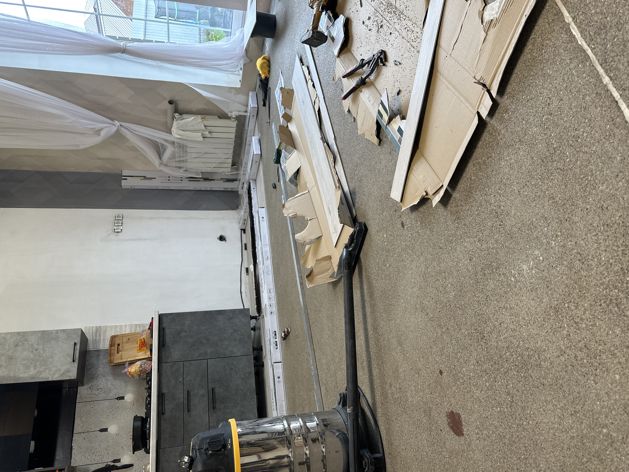 This is a photograph of an interior scene that appears to be under renovation or construction. The subjects include building materials such as long, flat pieces of wood or laminate flooring, cardboard boxes, and what looks like part of a disassembled kitchen cabinet. There is also a wet/dry vacuum cleaner in the foreground. The setting is a room with a concrete floor.
In the background, there is a window with sheer white curtains, a radiator, and what could be kitchen cabinets or built-in storage units. The walls are painted in neutral tones, with one section showing a geometric patterned wallpaper or tile. There are some tools visible on the floor, including pliers and a hammer. A yellow object, possibly a piece of safety equipment like a hard hat, is also present.
The overall impression is one of a work in progress, with materials and tools scattered as if someone was actively working on a project. There are no people visible in the image. The lighting suggests it is daytime, but the weather cannot be determined.
There are no prominent text elements visible in the image. The location is identified as Kuyuki, Russia.