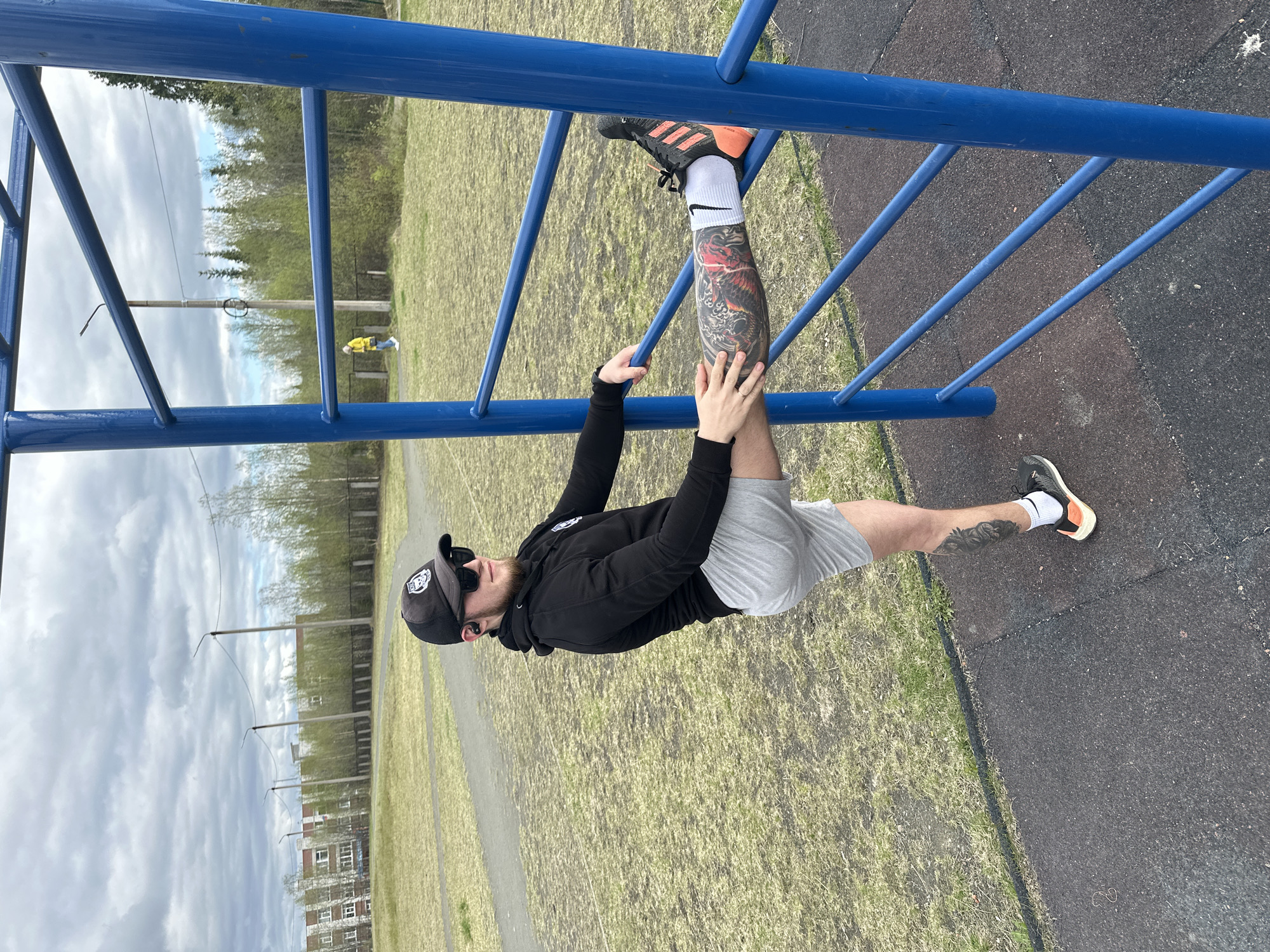 A man wearing a black cap, sunglasses, a black long-sleeved shirt, and grey shorts is suspended upside down from a blue metal jungle gym. He has tattoos on his lower left leg and is wearing a black and orange sneaker on his right foot and a white sock with a black and orange sneaker on his left foot. He is gripping the bars of the jungle gym with both hands. The jungle gym is situated on a playground surface, and beyond it is a grassy area with sparse vegetation, trees, and buildings in the background. The sky is overcast. The location is in the city of Unknown, Unknown.