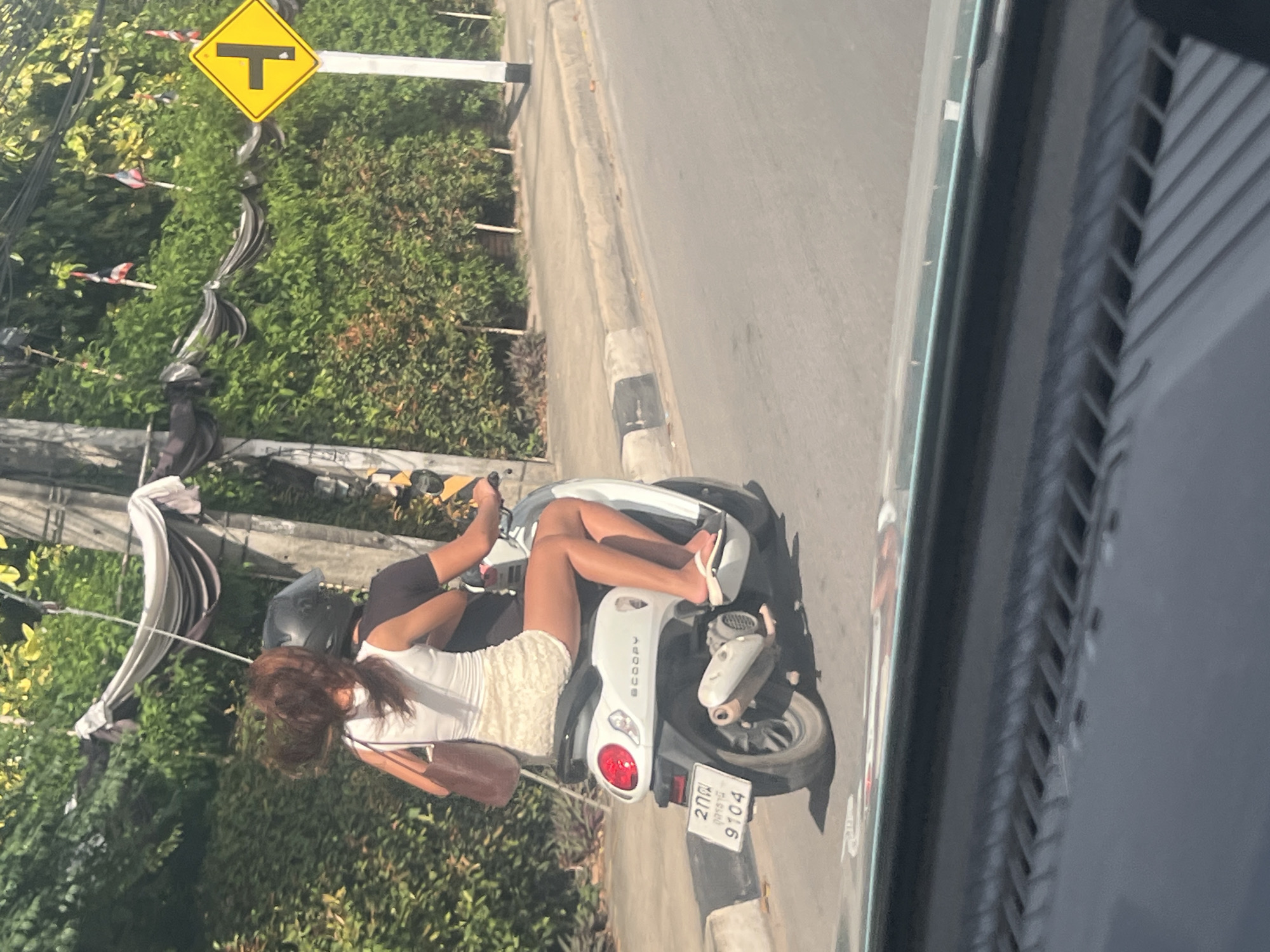 Two individuals are riding a white scooter on a paved road in a tropical environment. The person in front is wearing a black helmet and a dark t-shirt. The person behind, a woman with long brown hair, is wearing a white sleeveless top and a light-colored, textured skirt. She is holding a brown purse on her left shoulder and has her arm around the person in front. Both are wearing casual footwear. The scooter has a visible license plate with Thai characters and the number "9104." The surroundings include lush green foliage, trees, and a sidewalk with a concrete curb. A yellow diamond-shaped traffic sign with a T-intersection symbol is visible on a pole to the right. The sky is bright, suggesting daylight. The scene appears to be captured from inside a vehicle, as indicated by the blurry foreground of a dashboard and windshield. This location is identified as Ko Samui District, Thailand.