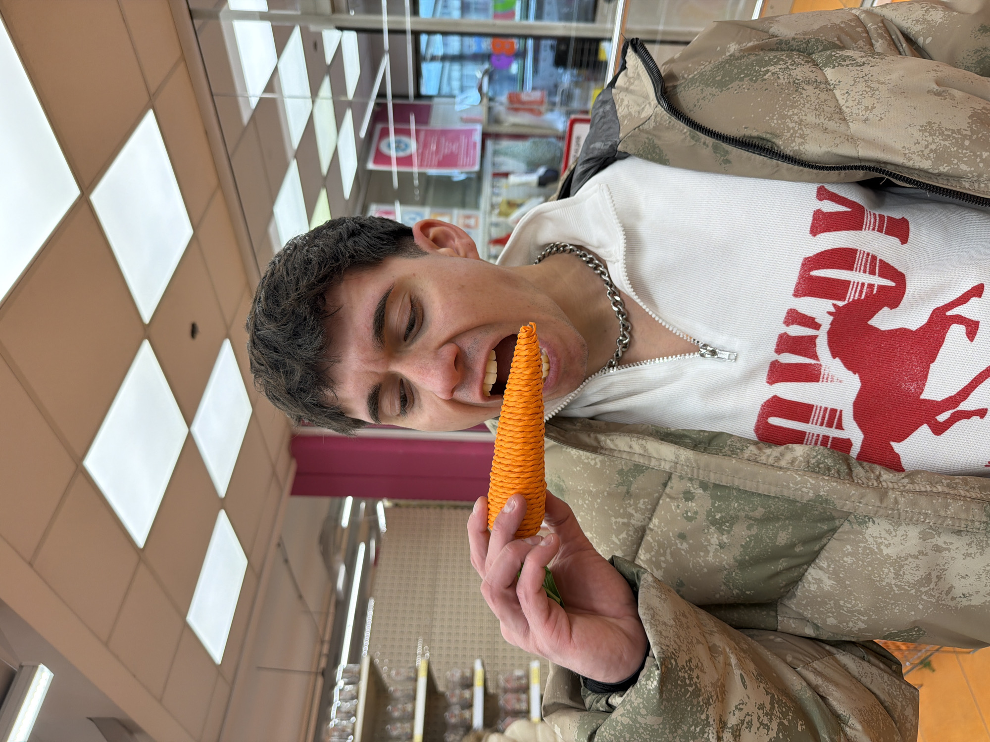 A man is indoors, holding an orange, carrot-shaped object in his right hand and partially putting it into his mouth. The man has short, dark hair and is wearing a silver chain necklace, a white zipped top with a red graphic design, and a camouflage pattern jacket. The location appears to be inside a store with fluorescent lights and shelves of items visible in the background. The city of Sofia, Bulgaria, is not discernable in the image.