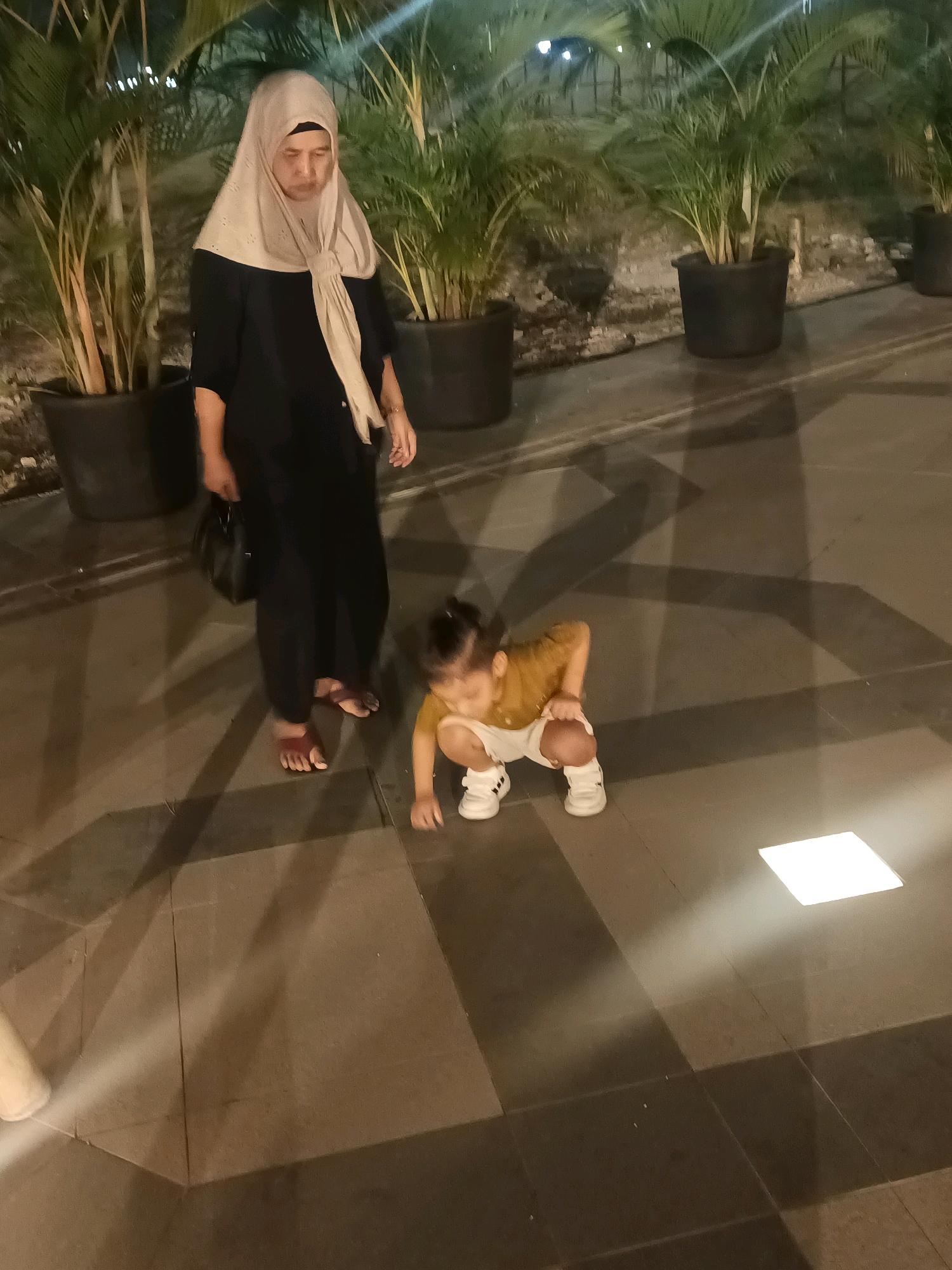 In the city of North Jakarta, Indonesia, two individuals are present on a paved walkway. A woman, wearing a black abaya and a beige hijab, stands to the left. Her head is bowed, and she appears to be looking downwards. In front of her, a young child is squatting, dressed in a mustard-colored shirt and white shorts. The child's hands are resting on their knees. The walkway is paved with large square tiles in varying shades of grey and brown, with shadows cast across the surface. In the background, several large potted plants with palm-like fronds are visible. A square-shaped light fixture is embedded in the walkway to the right of the child, emitting a bright glow. The overall lighting suggests it is evening or night.