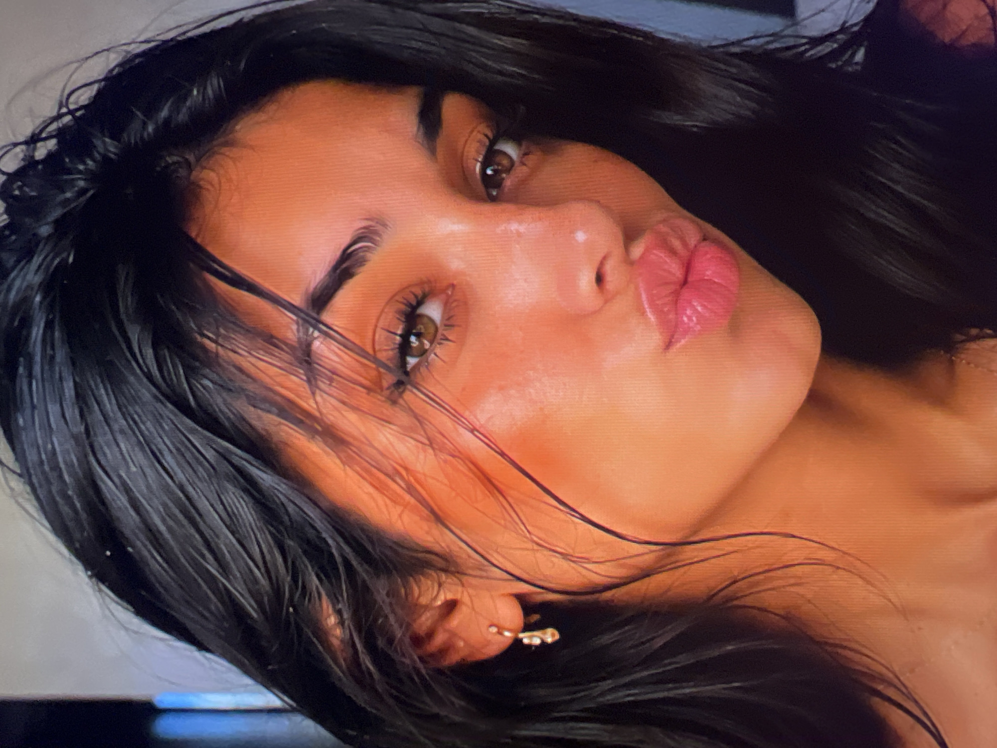 The image is a close-up, head-and-shoulders shot of a young woman with dark, wet hair and warm-toned skin. She is wearing a delicate gold earring with a dangling charm. Her lips are pursed in a kiss pose, and she has on a glossy pink lipstick. Her eyes are a light brown, and her eyebrows are well-defined. The background is blurred, but appears to be an indoor setting with some vertical lines suggesting shelving or a wall. There are no discernible features that place the location in Athens, Greece.