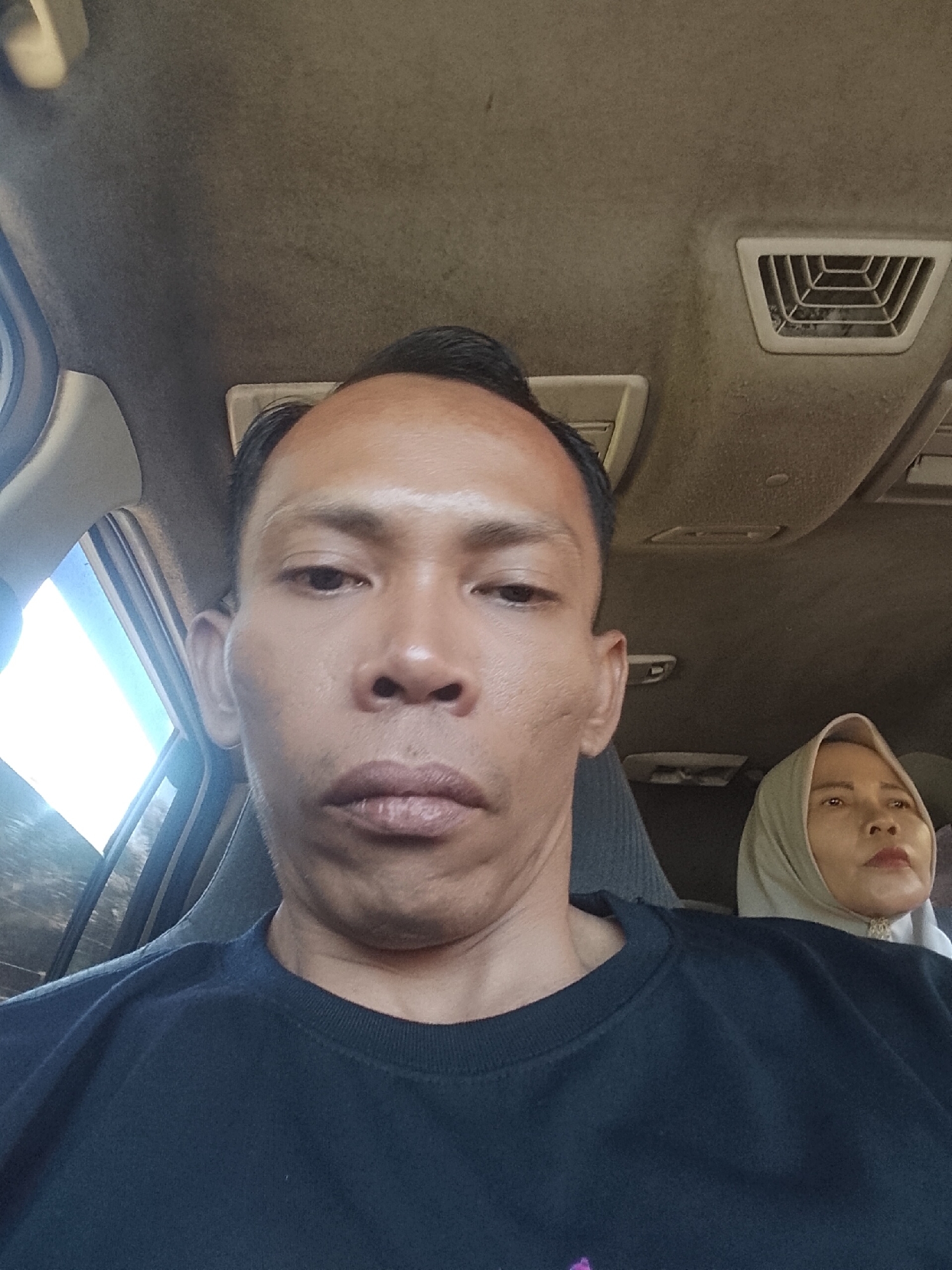 A selfie taken from inside a vehicle shows a man in the foreground, his face close to the camera. He has short, dark hair, dark eyes, and light skin. He is wearing a dark blue t-shirt. Behind him, a woman is visible, wearing a light-colored hijab. She is looking to her right. The interior of the vehicle is visible, with an air vent on the ceiling and part of the dashboard. The background appears to be outdoors, with sunlight streaming in from the left. The image appears to have been taken in Tiang Tara, Indonesia.