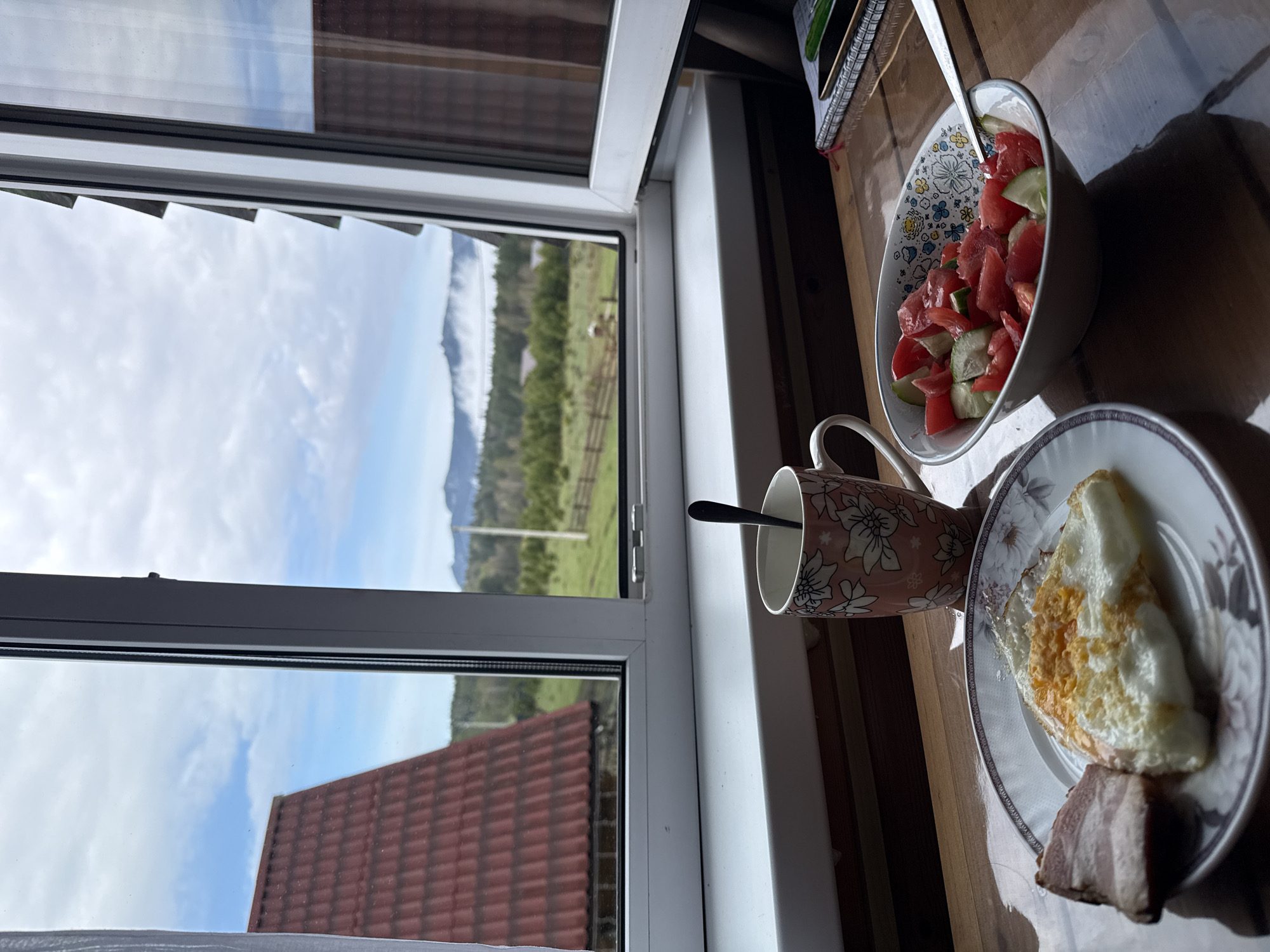 The image depicts a breakfast setting indoors, likely near a window, with a view of the outdoors. The breakfast consists of a plate with a fried egg and a piece of meat, a bowl of tomato and cucumber salad, and a mug with a spoon inside. Outside the window, there's a view of green fields, a fence, some trees, and mountains in the distance under a cloudy sky. A red tiled roof is visible in the lower part of the window. The scene could be in Tyulyuk, Russia, given the landscape.