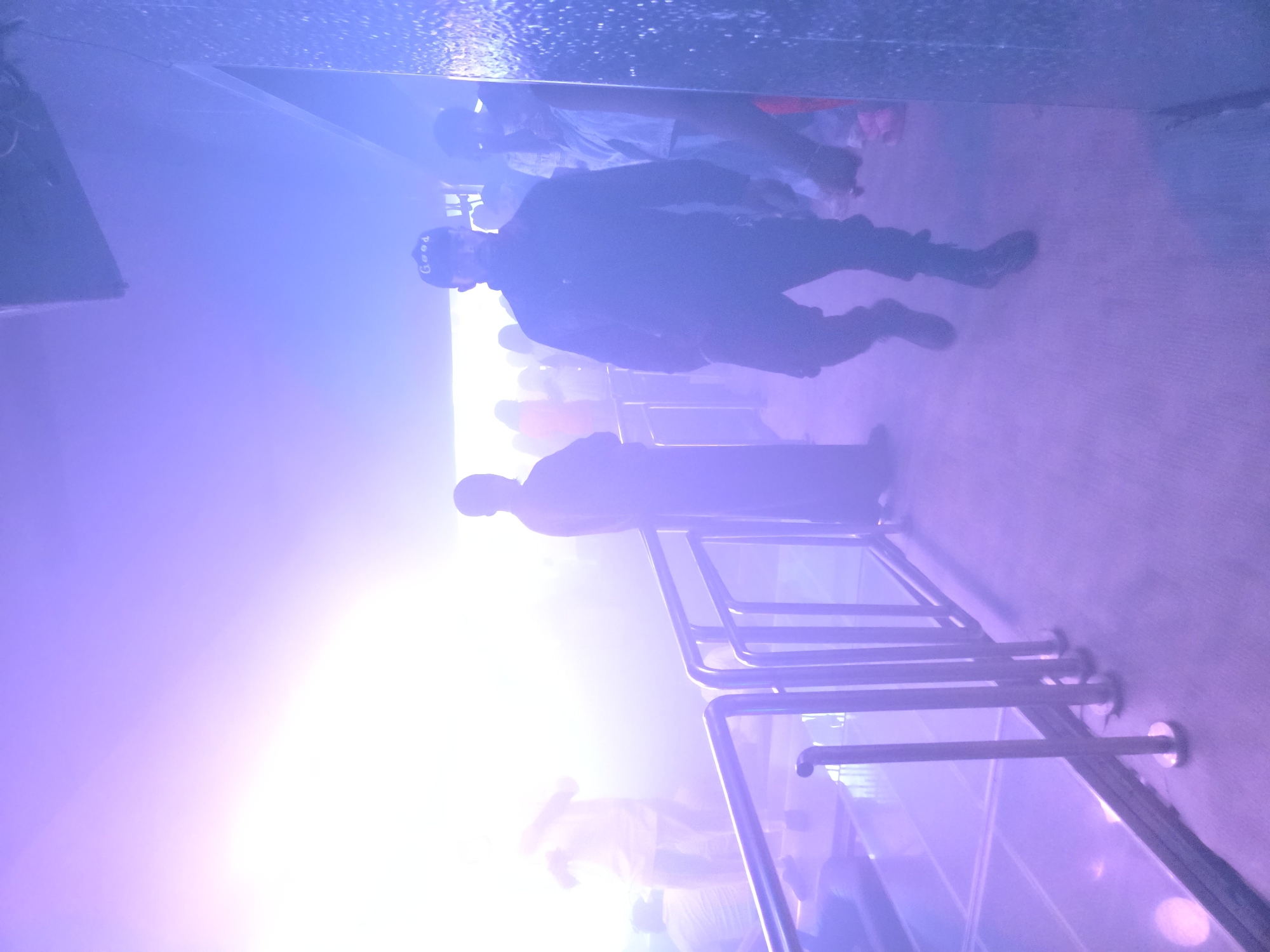 An interior shot shows a group of people standing in a dimly lit venue, possibly a nightclub or concert hall, with purple and pink lighting casting a hazy glow. In the foreground, a person wearing a black cap with "Good" written on it and black clothing walks towards the viewer. Behind them, other individuals are visible, some blurred by the lighting and atmosphere. A metal railing runs along the left side of the frame, separating a slightly elevated, reflective floor from the main walkway. The overall impression is one of a lively, possibly crowded, entertainment space. The location is Gura Top, Nigeria.