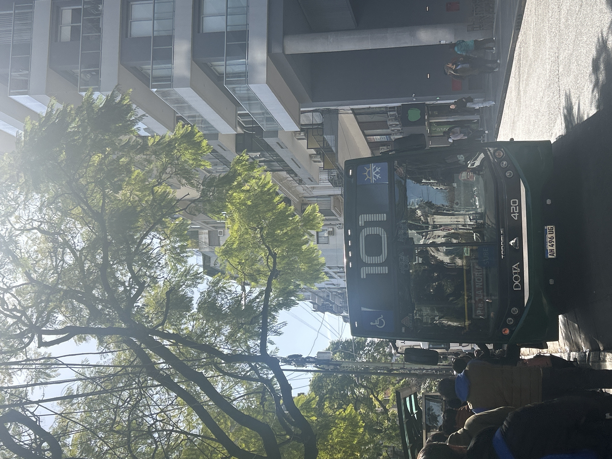 The image captures a street scene in Buenos Aires, Argentina, likely during daylight hours given the bright sunlight and shadows. The foreground is dominated by the front of a green public bus, with the number "101" prominently displayed on its windshield. The bus has a "DOTA" logo and the number "420" visible below the windshield. There's a license plate with the characters "AH 496 UG". A blue wheelchair accessibility symbol is also present on the front of the bus.
To the left of the bus, a group of people are gathered, appearing to be waiting or boarding. Some are wearing casual clothing, including a person in a blue jacket and another in a brown vest. The bus is stopped at what appears to be a bus stop, with a covered shelter visible behind the waiting passengers.
The background reveals a typical urban environment. To the right, a modern multi-story building with balconies and glass railings stands tall. Trees with green foliage, partially obscuring the sky, line the street, casting shadows on the pavement. Other buildings and utility poles are visible further down the street, contributing to the urban landscape. The overall impression is a snapshot of daily public transportation in a bustling city.