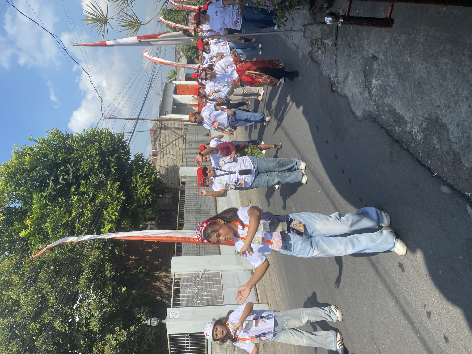 A group of people, mostly women and girls, are walking in a parade down a paved street in Kediri City, Indonesia. Many participants are wearing white t-shirts and red headbands, and some are wearing red hats. Several individuals have sashes or necklaces made of what appear to be money. Indonesian flags are prominently displayed, with red and white banners attached to poles along the street. The buildings lining the street are simple, with brick walls and tiled roofs. The sky is blue with scattered clouds, indicating it is a sunny day. The street shows signs of wear and tear, with cracks and patches in the asphalt. Some of the people are gesturing or smiling, suggesting a festive or celebratory atmosphere.