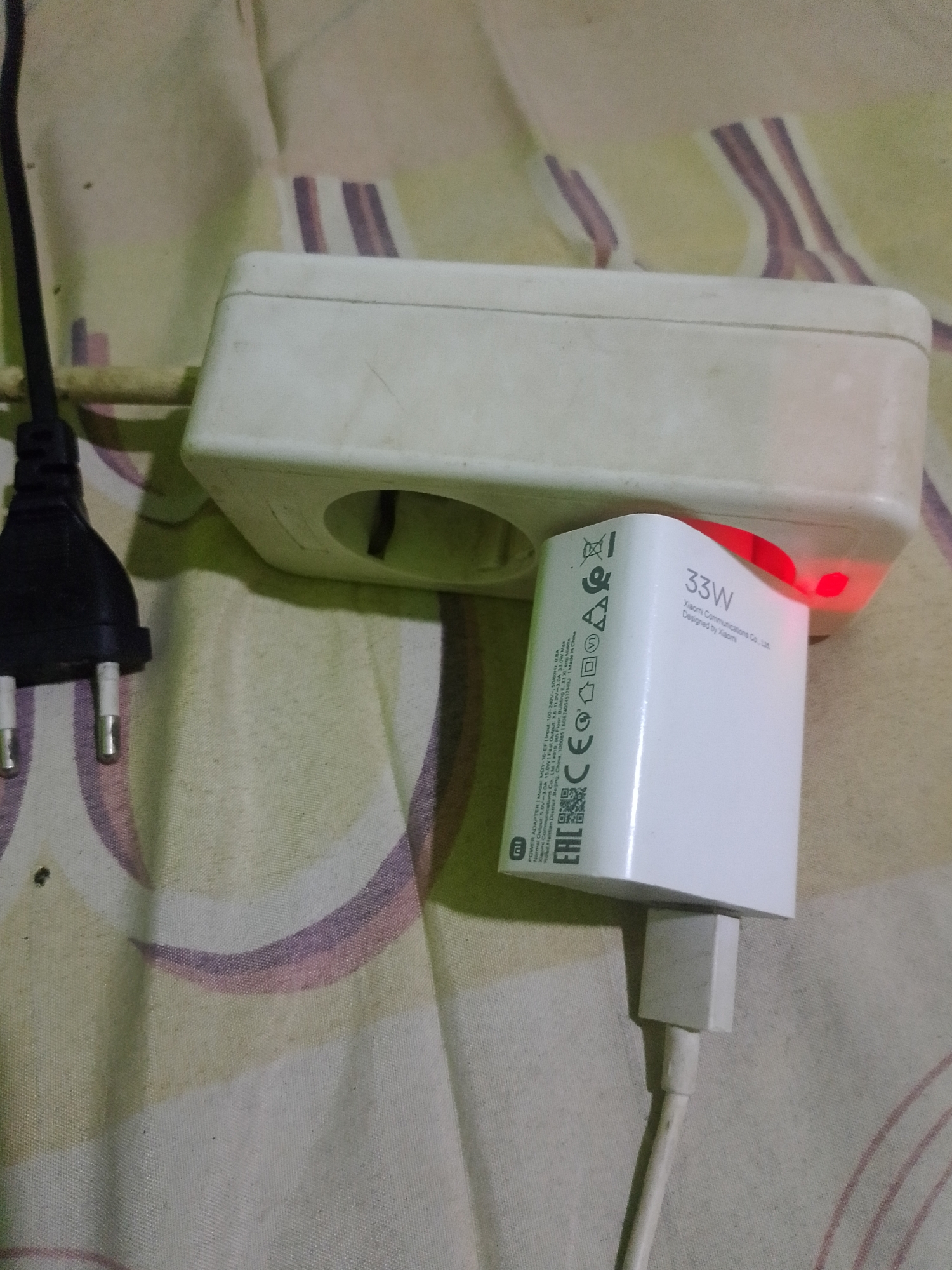 The image shows a close-up view of a white power adapter plugged into a white power strip. The power adapter has "33W" printed on it in large letters and "Xiaomi Communications Co., Ltd. Designed by Xiaomi" printed below. It also has several certifications and labels, including "CE", "EAC", and "MI". The power adapter has a red indicator light that is illuminated. A black electrical plug is also visible, partially obscuring the power strip. The objects are situated on a patterned fabric surface, likely a bed or couch, with a light green background and stylized purple and pink stripes. The scene is indoors, with no indication of the specific location within Banyuwangi, Indonesia, other than it is a residential or commercial indoor setting. There are no people visible in the image. The image is taken from a slightly elevated angle, looking down on the objects.