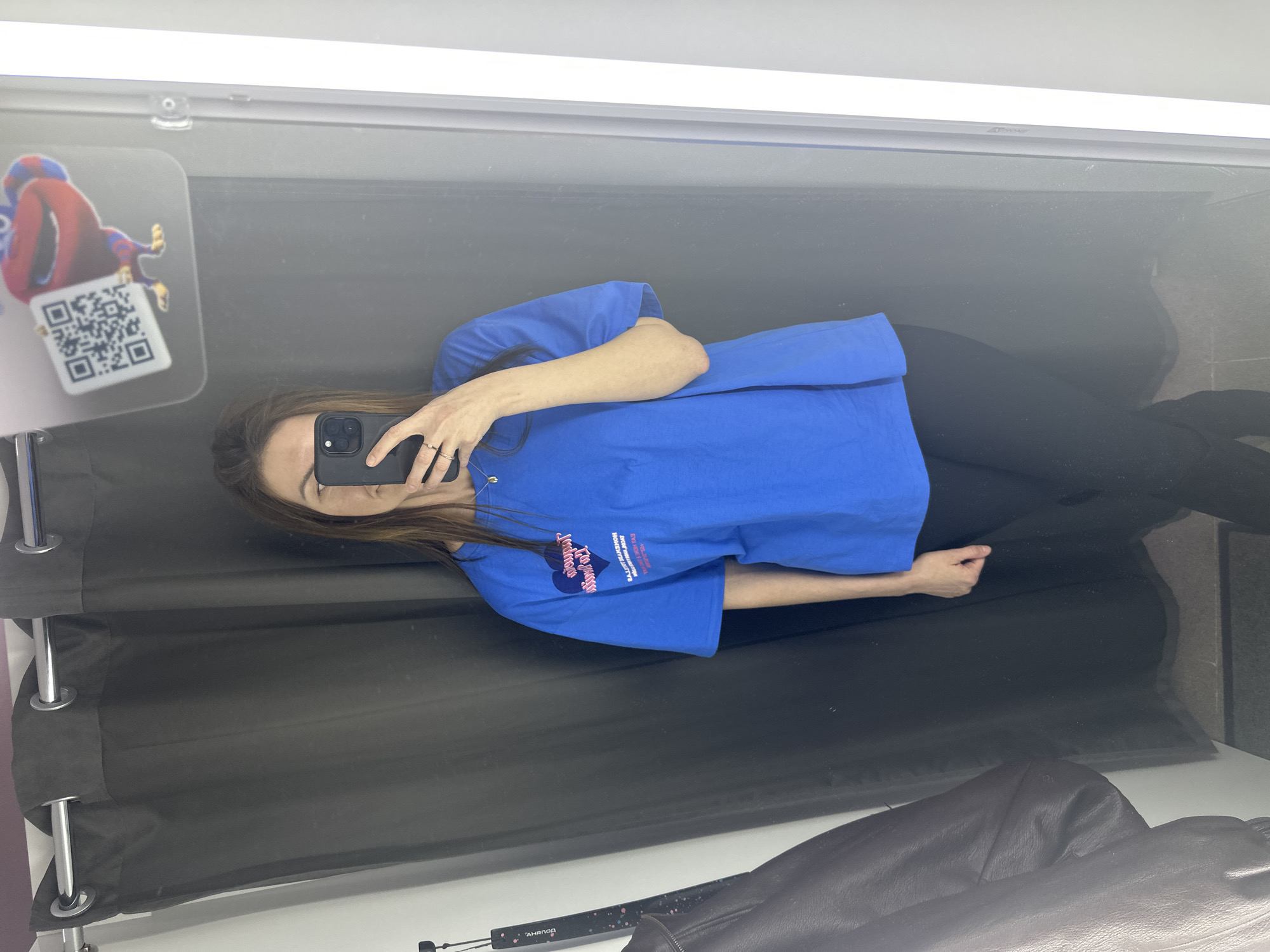 The image, captured in Chaikovsky, Russia, displays a reflection of a woman taking a mirror selfie in what appears to be an indoor setting, likely a fitting room. The woman has long brown hair and is wearing a royal blue short-sleeved t-shirt and black pants. She holds a dark-colored smartphone to her face, partially obscuring it, with her left hand visible, adorned with rings.
The blue t-shirt features a graphic design on the front. Clearly visible text includes "LA MOVIDA," and within a heart shape, "BALENCIAGA MAISON FONDÉE EN 1917 PARIS," along with "DELUXE" and other smaller, unreadable text.
To the upper left of the reflection, a clear plastic display hangs, showing a colorful, cartoon-like figure in red and blue, along with a prominent QR code. The setting includes dark gray or black fabric panels, possibly privacy curtains, with silver grommets and rods on the left side of the reflection. Bright artificial lighting fixtures are visible along the top edge of the mirrored surface. A dark brown or black jacket is partially reflected in the bottom right corner of the image. The scene is illuminated by artificial light, and no specific time of day or weather conditions are discernible.