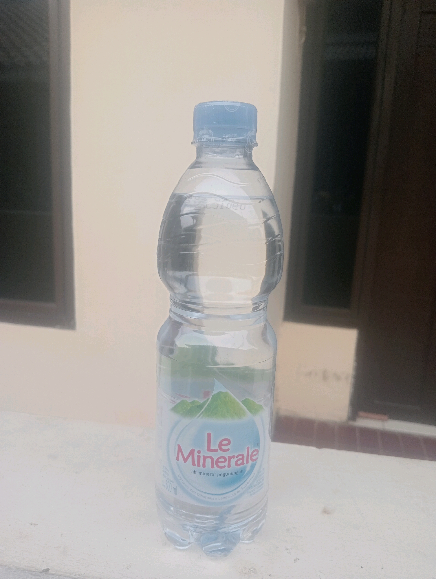 A medium shot shows a clear plastic bottle of Le Minerale bottled water, filled with water and sealed with a light blue cap. The bottle is positioned centrally on a light-colored ledge or windowsill, with a textured surface. Behind the bottle, the background consists of a neutral-colored wall, possibly light beige or cream, with two dark rectangular shapes that appear to be windows or openings with dark frames. To the right of the wall, a darker section suggests a doorway or a shadowed area. The lighting is soft and diffused, indicating an indoor or covered outdoor setting. There are no people present in the image. The overall scene is mundane and unremarkable. This image is not associated with the city of East Jakarta, Indonesia; it depicts a generic scene likely taken in a residential or commercial building.