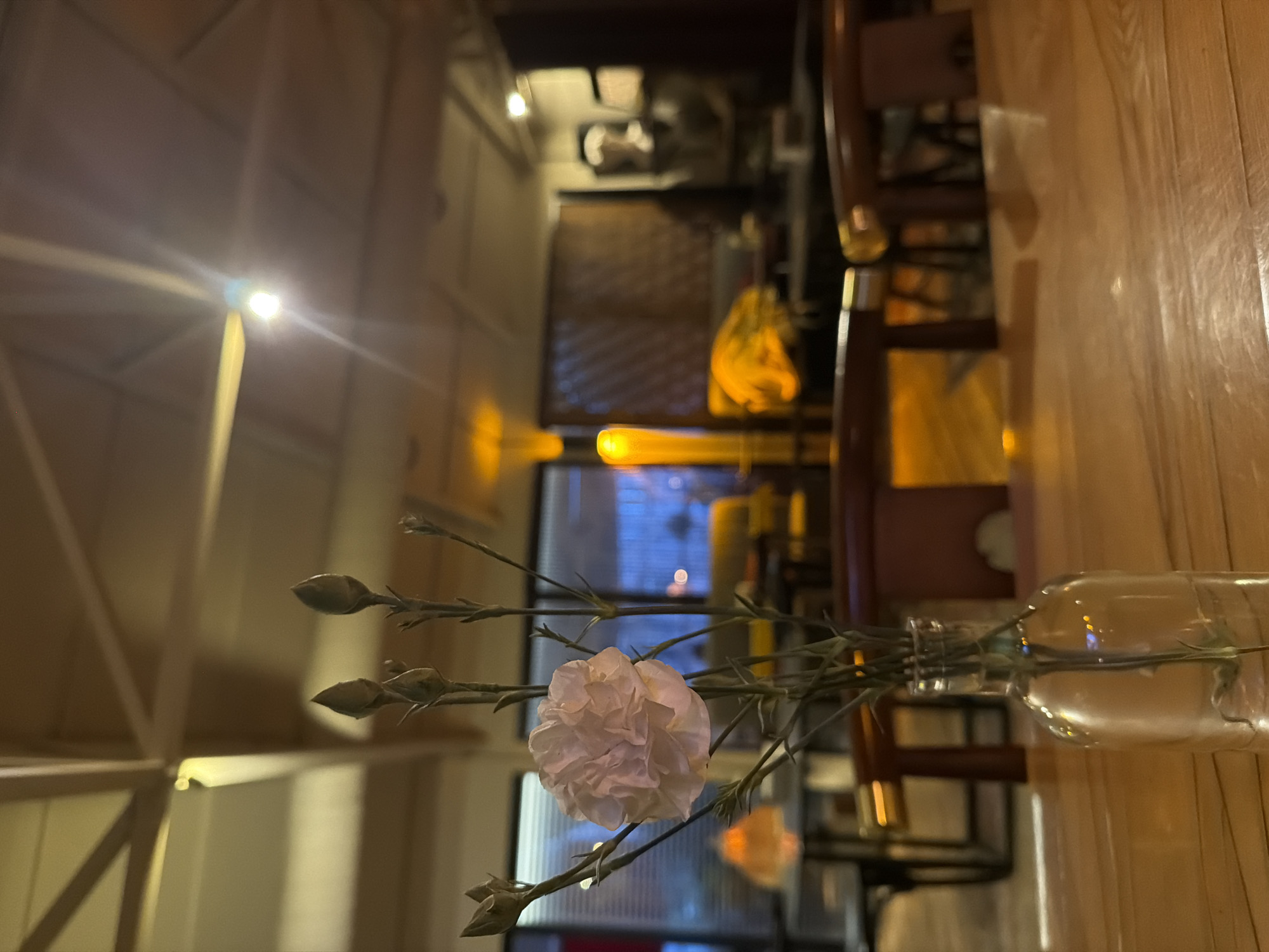 The image shows a blurry indoor scene. A white flower is in focus and placed in a glass bottle. The bottle sits on a wooden table. A chair is near the table. A curtain is hanging in the back. The location and the presence of people are indiscernible.