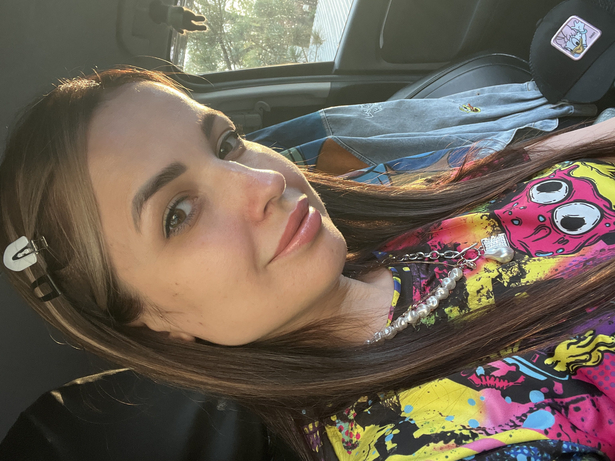 A medium shot depicts a young woman in a vehicle. The woman is positioned on her side, facing the camera. She has long, brown hair, fair skin, and light-colored eyes. She is wearing a colorful, patterned shirt. A pearl necklace and a silver chain are visible around her neck. A hair clip is fastened in her hair near her temple. Sunlight is striking her face. Behind her, a denim jacket and a baseball cap with a cartoon character emblem are visible. The background includes the car's interior, a sunroof, and foliage visible through the sunroof. The location is Unknown, Unknown.