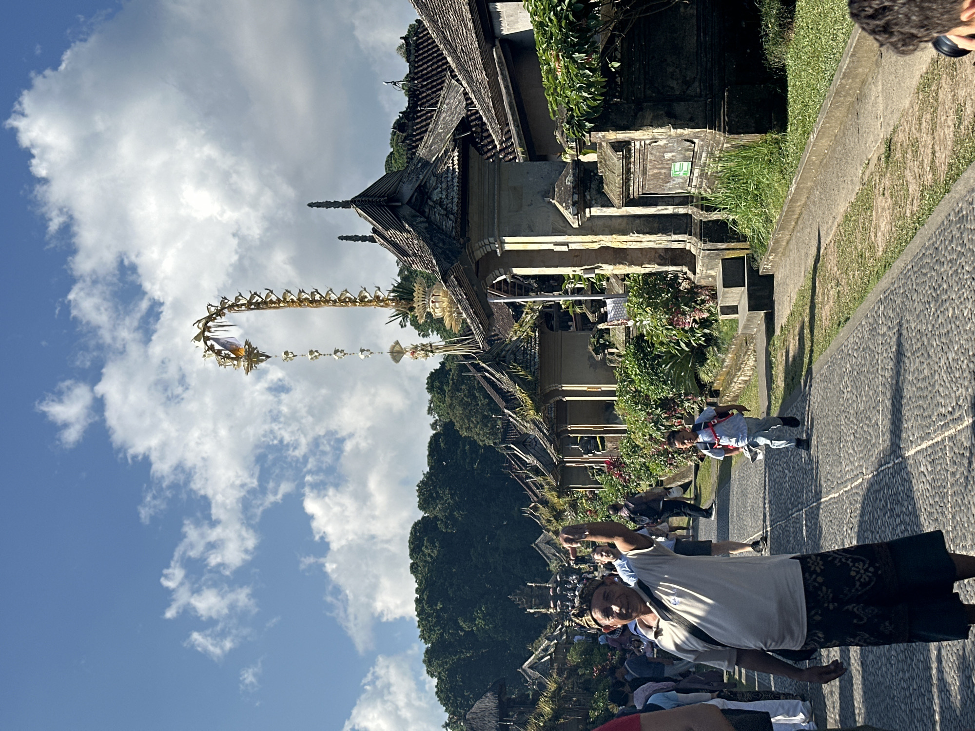 The image depicts a ceremonial procession in the city of Bangli, Indonesia, likely at the Pura Kehen temple complex, given the traditional Balinese architecture and the presence of a towering, ornate "penjor" (ceremonial bamboo pole). In the foreground, an older man with a patterned sarong and a white shirt is looking towards the camera with his left arm raised. To his right, a young boy in a blue shirt and backpack is running down a cobblestone path. The path is lined with traditional Balinese buildings featuring intricate stone carvings and thatched roofs. Lush green vegetation grows along the sides of the path. In the background, a dense green forest rises up a hillside under a bright blue sky with scattered white clouds. The atmosphere appears festive and communal, with other people visible in the middle ground and background, suggesting a public event or celebration.