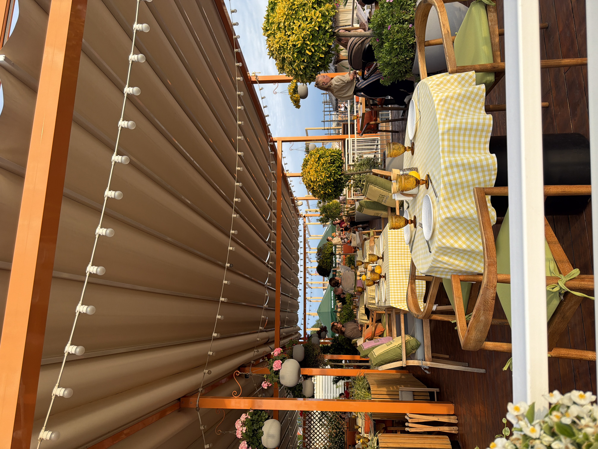 This media file depicts an outdoor seating area of a restaurant or cafe, likely in Sochi, Russia, during the daytime. The scene is set under a beige retractable awning, supported by orange metal beams. Strings of white globe lights are strung along the awning's underside, suggesting it could be an evening setting, though the bright sky indicates daytime.
Several tables are set for dining, covered with yellow and white checkered tablecloths. Each table is adorned with amber-colored glasses and white plates. Wooden chairs with green cushions are arranged around the tables.
In the background, there are lush green trees with yellow foliage, adding to the vibrant atmosphere. Some people are visible seated at tables, engaged in dining and conversation, though their faces are not clearly discernible. The overall mood appears relaxed and leisurely, indicative of a pleasant day out.
The foreground features hanging planters with pink flowers and white decorative lanterns, adding a touch of charm to the outdoor space. The wooden decking flooring and the style of the outdoor furniture suggest a seaside or a resort-like location. There is no visible text in the media file.