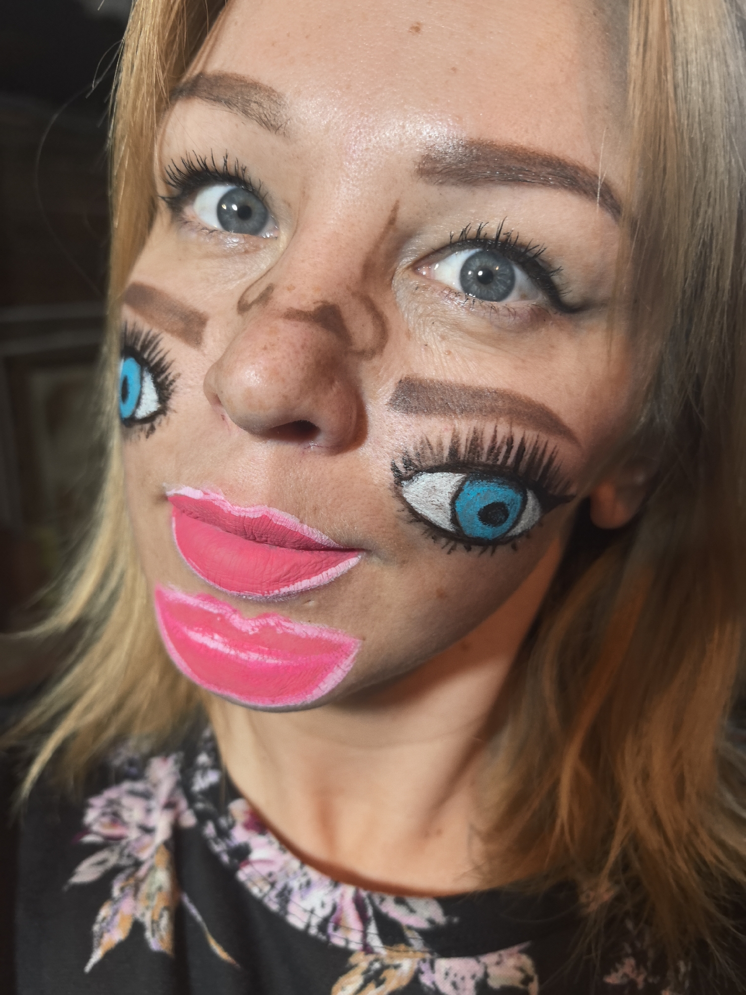 A close-up portrait shot in Noviy, Russia, features a woman with blonde or light brown hair adorned with elaborate, artistic makeup that creates a surreal, distorted facial effect. Her natural right eye, a light blue-grey, is wide open and gazes directly at the viewer, framed by dark mascara on her lashes and a neatly shaped eyebrow. Her nose is contoured with darker makeup lines, giving it a stylized, almost caricatured appearance.
On the left side of her face, a large, exaggerated eye is painted on her cheek, featuring a vibrant blue iris, a black pupil, and long black eyelashes, with a brown eyebrow painted above it. Below her natural mouth area, two sets of bright pink lips are painted onto her lower face. The upper set of painted lips appears as her main mouth, slightly parted, while a smaller, similar pair of lips is painted below it. Both sets of painted lips are outlined in white.
She is wearing a dark top with a floral pattern in shades of pink, purple, and orange visible around her shoulder and chest. Her skin has some freckles visible. The background is dark and out of focus, suggesting an indoor setting. The lighting is bright and direct, highlighting the details of her makeup. No visible text is present in the image.