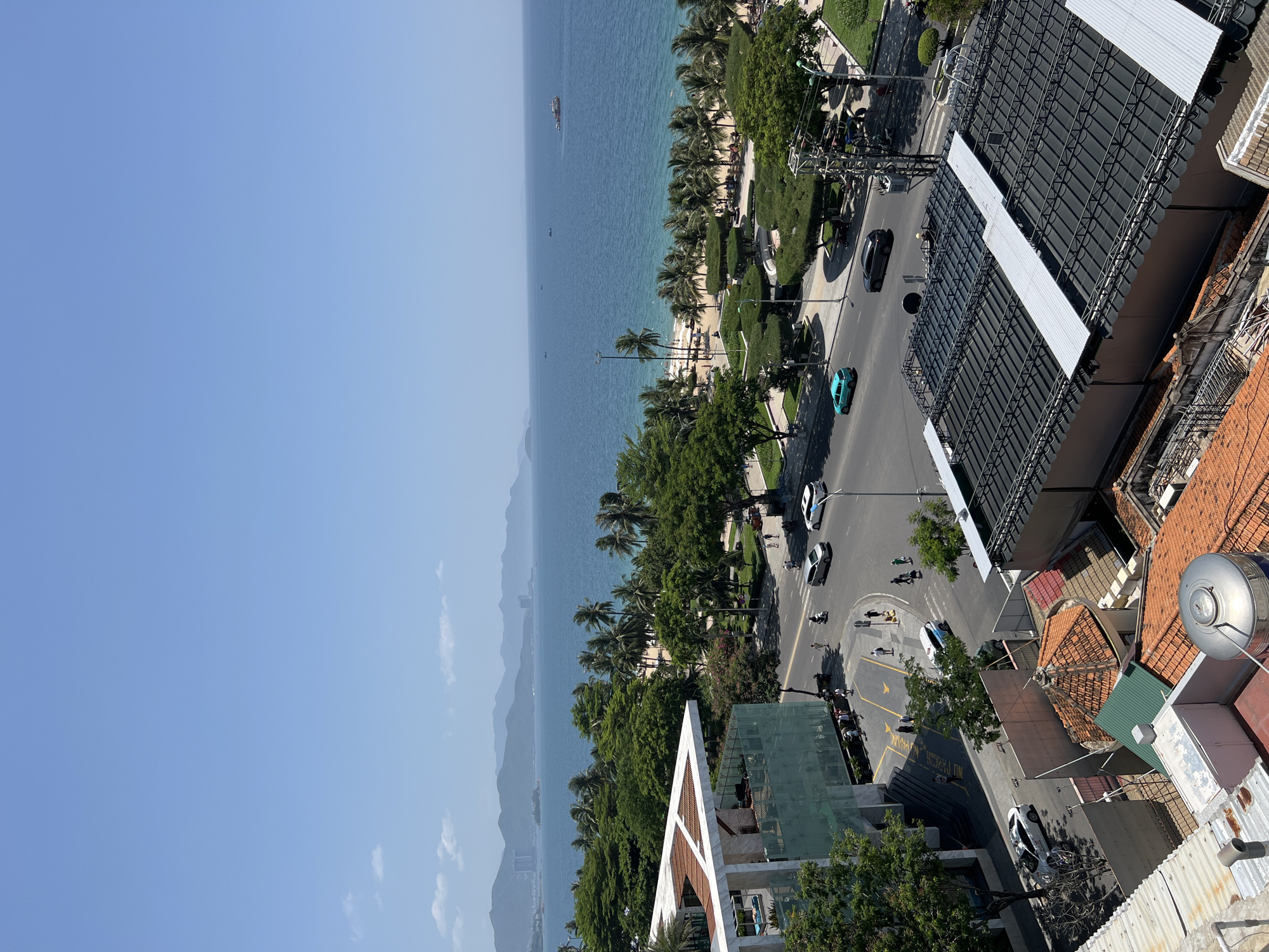The image is a high-angle, eye-level view of a coastal city street in Nha Trang, Vietnam. The scene is dominated by a bright, clear blue sky with a few scattered white clouds. In the distance, a range of hazy mountains stretches across the horizon.
The middle ground features a wide, paved road with several cars, a scooter, and pedestrians. A turquoise car is driving on the road, alongside a black car and a white car. People are walking on the sidewalks and crossing the street.
Adjacent to the road is a palm-lined beach with golden sand. The ocean is a deep blue, with a few small boats visible in the distance. Several larger boats, including a cargo ship, are further out at sea. The beach is dotted with palm trees and some beachgoers.
In the foreground, a modern building with a white, angular roof is partially visible on the left. The building has large glass windows, and scaffolding is present on one side, indicating ongoing construction or renovation. On the right, the rooftops of several buildings are visible, characterized by orange tile roofs and some modern structures with metal roofing.
The overall impression is a sunny day in a bustling coastal city, likely a tourist destination, with a mix of modern architecture and natural coastal scenery.