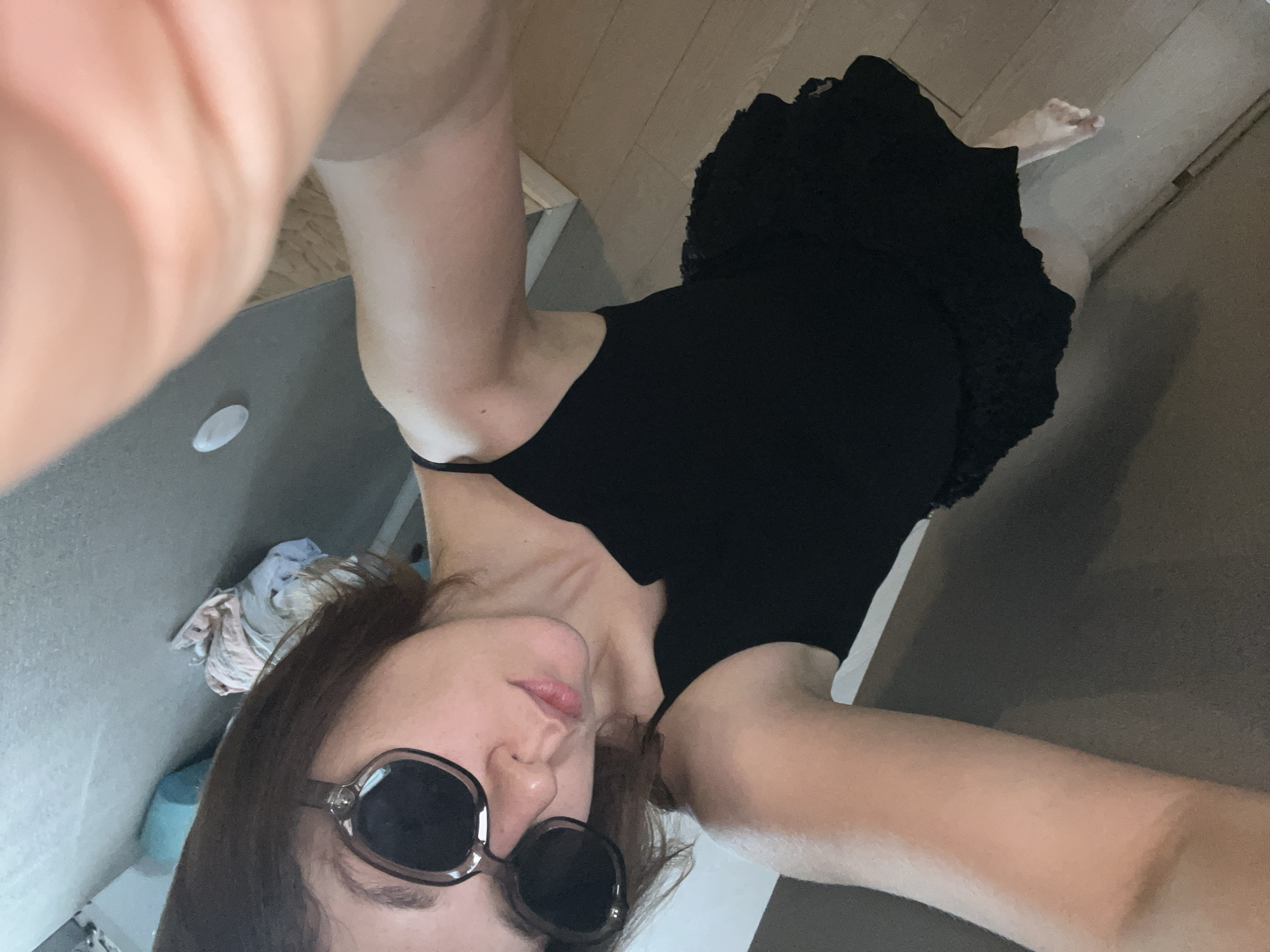 A woman is taking a selfie. She is wearing sunglasses and a black dress. The setting appears to be indoors, with wooden flooring and a gray wall in the background. A pile of clothes is visible on a shelf or surface against the wall. The image does not provide any specific details that would indicate its location in Krasnoyarsk, Russia.